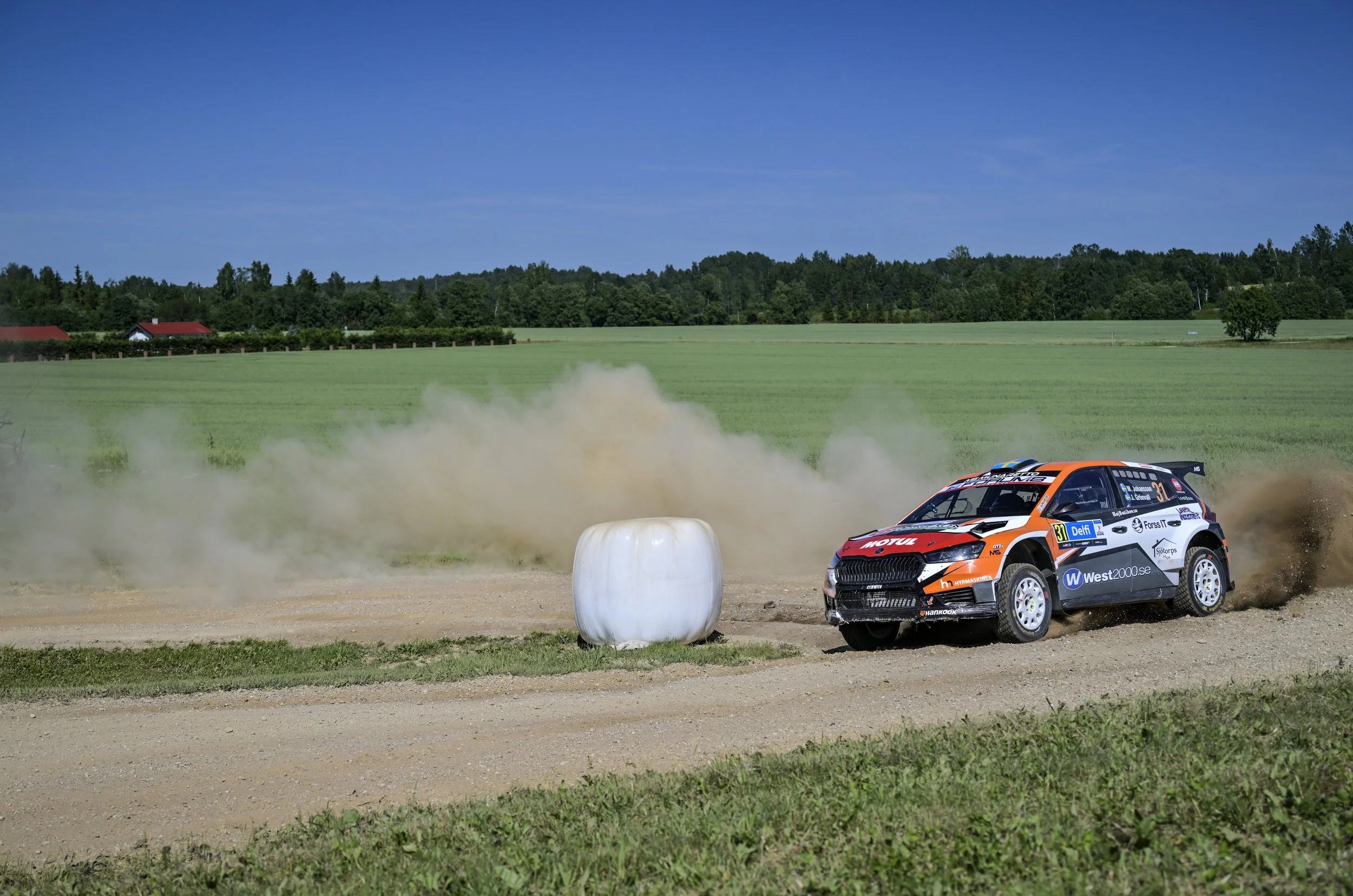 A rally car on a dirt track kicking up dust, with a rural landscape of green fields, trees, and houses under a blue sky in the background.