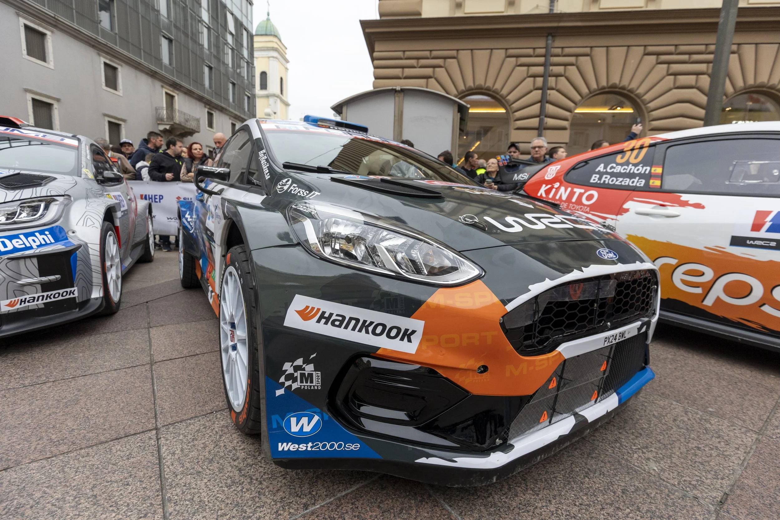 Several race cars are displayed outdoors with people gathered around, including a gray and orange Ford car in the foreground, a black and white car on the left, and a red and white car on the right, at an event with buildings in the background.
