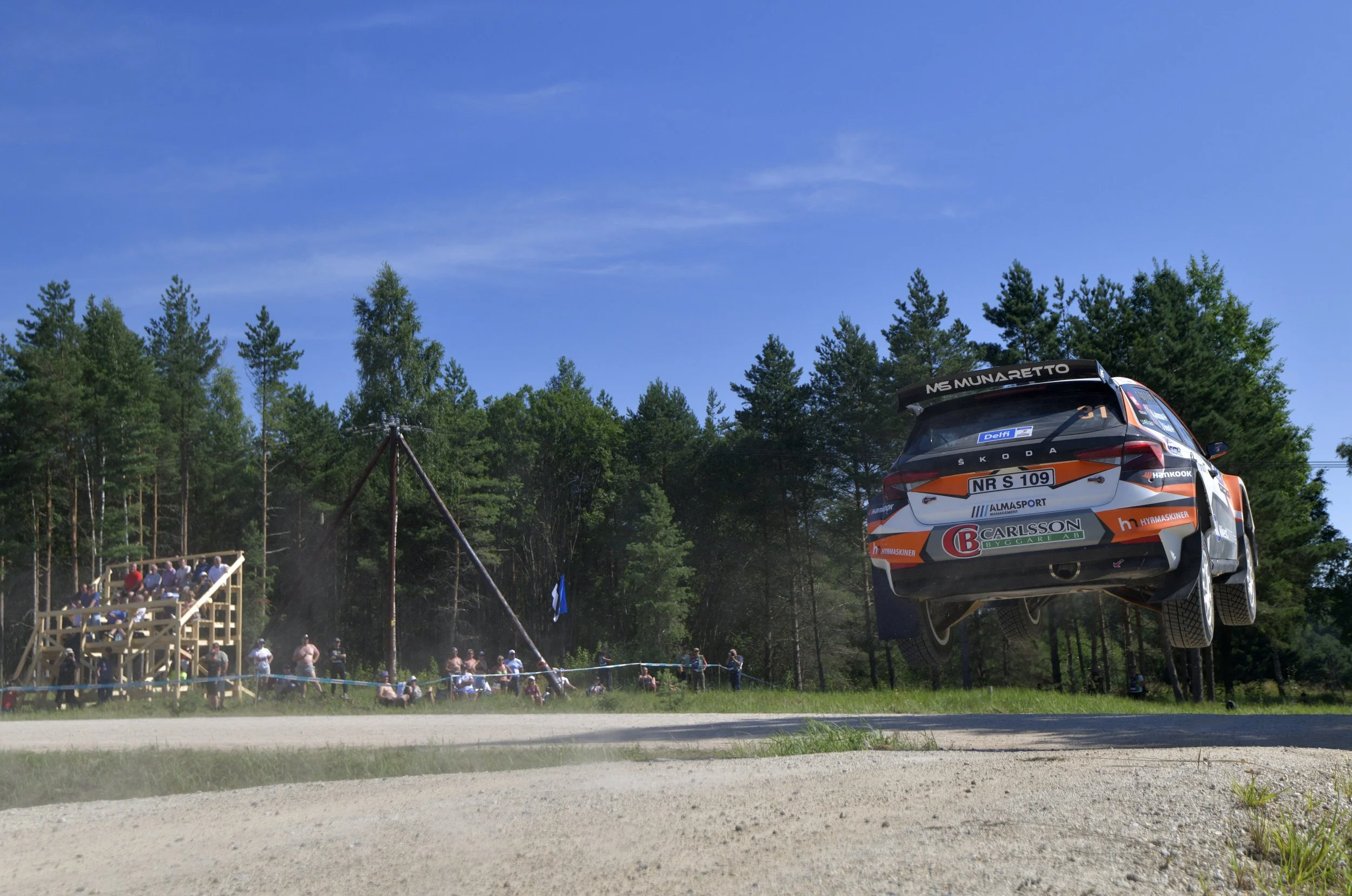 A rally car, number 31, is airborne during a race, with spectators watching from a wooden stand and forested area in the background on a clear day.