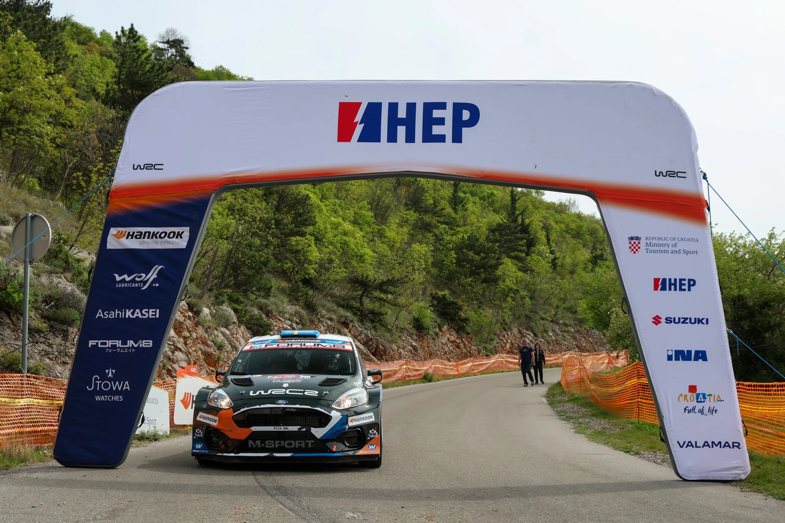 A racing car crossing a finish line under an arch with sponsorship logos during a rally event, surrounded by green trees and spectators.