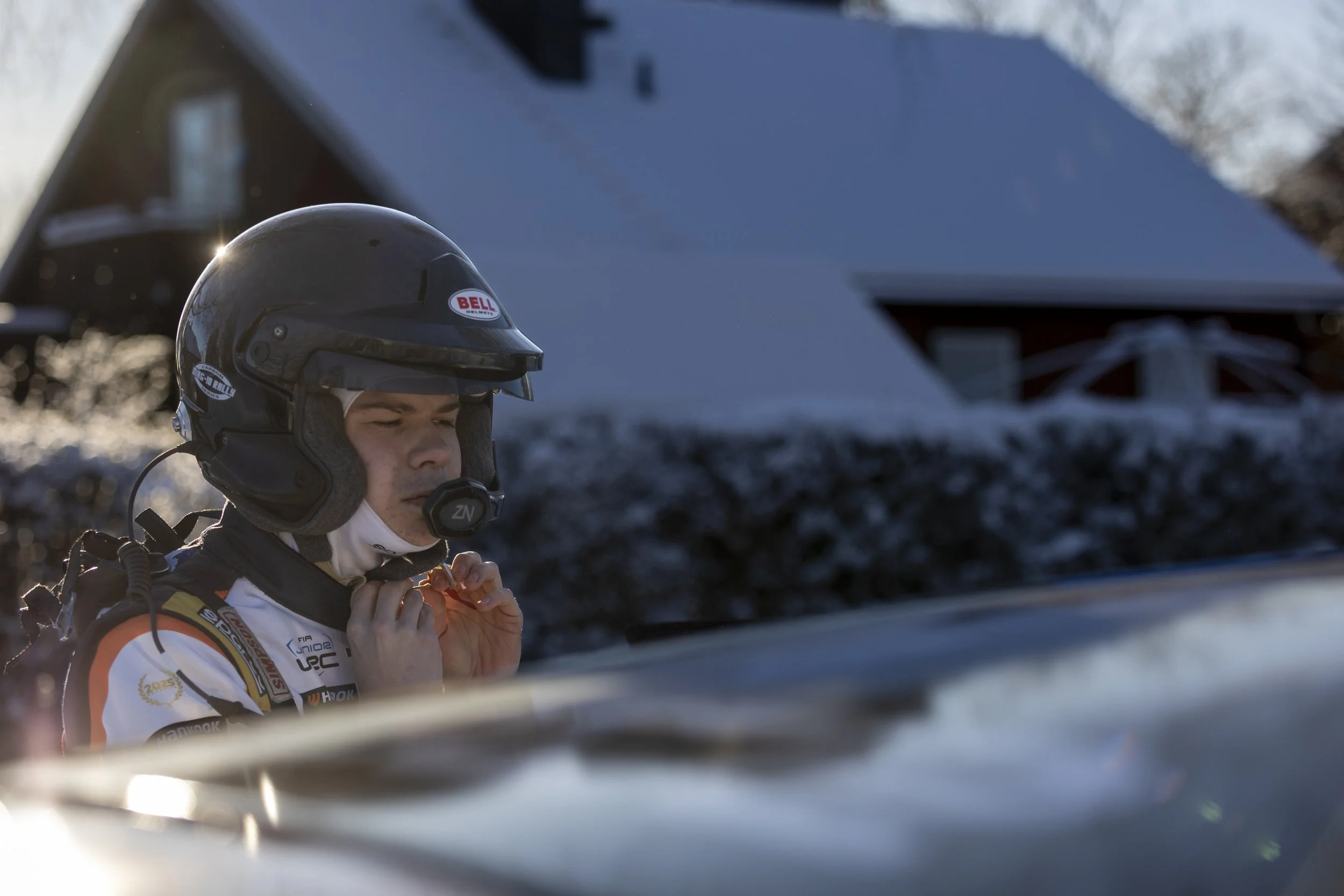 A man wearing a racing helmet and a racing suit, adjusting his seatbelt, standing outdoors with a building and trees in the background.