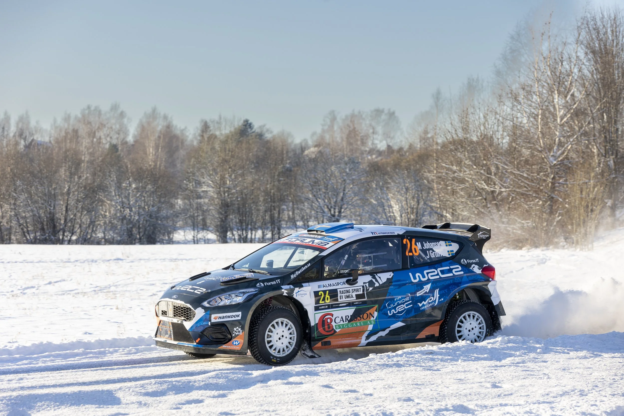 A rally car racing on snow-covered terrain during winter, with trees and a clear blue sky in the background.