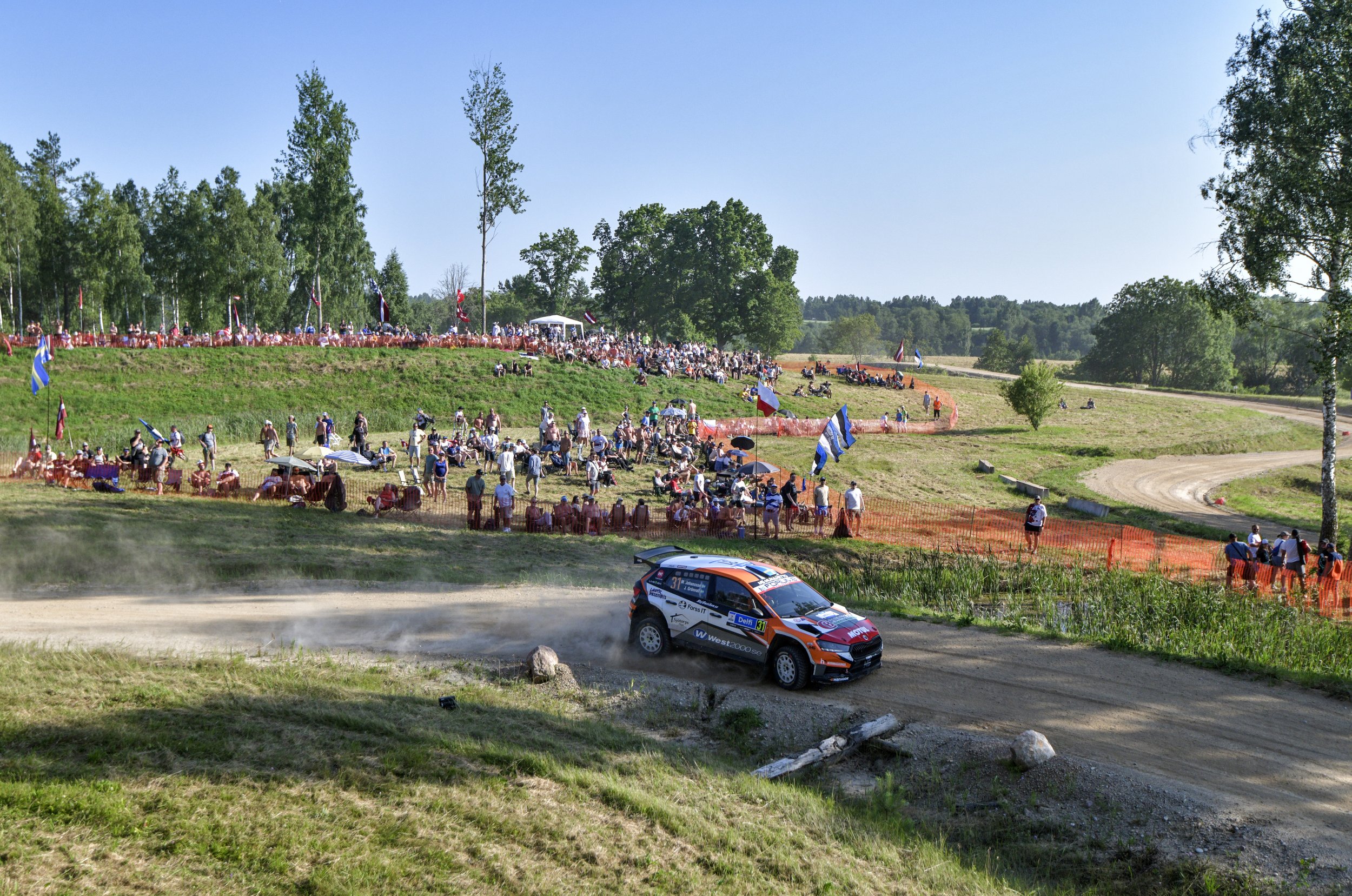 A rally car speeds along a dirt track with a crowd of spectators watching on a hillside, some under umbrellas, surrounded by trees and a bright blue sky.