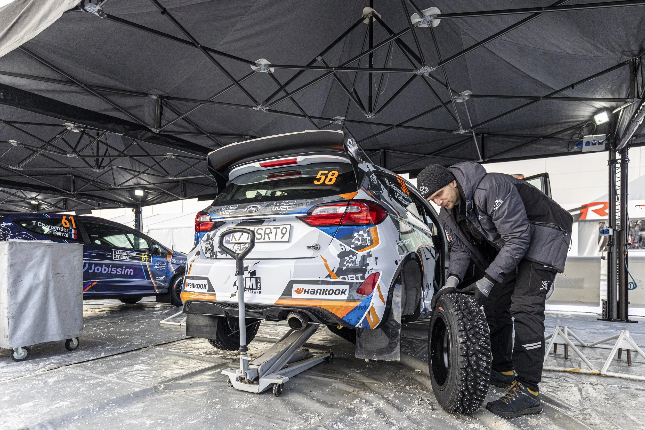A man working on a rally car inside a black canopy tent at a racing event. He is kneeling and using tools on the car, which has racing decals and sponsor logos.