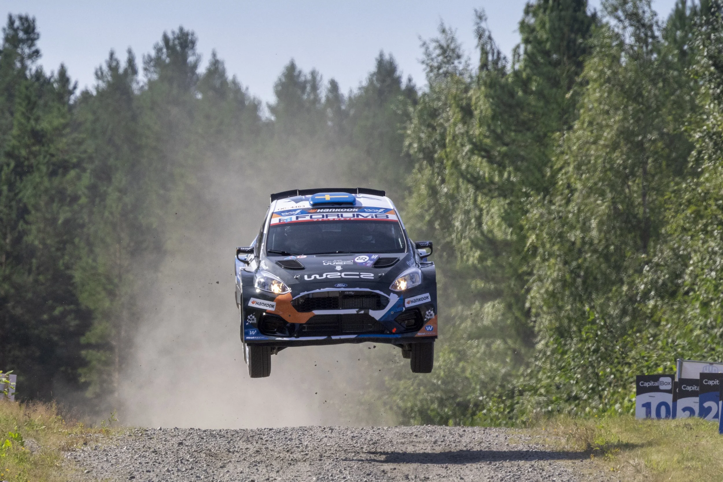 A rally car mid-air during a jump on a gravel road surrounded by green trees.