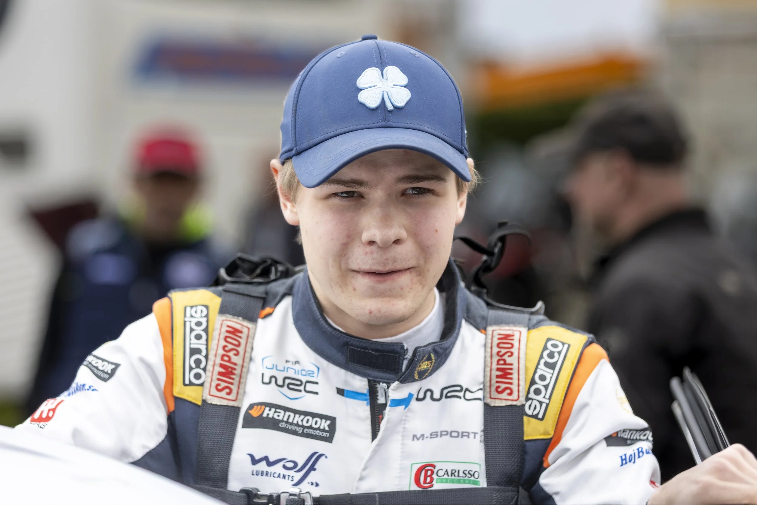 A young race car driver in racing gear, wearing a blue cap with a four-leaf clover patch, surrounded by people at a racing event.