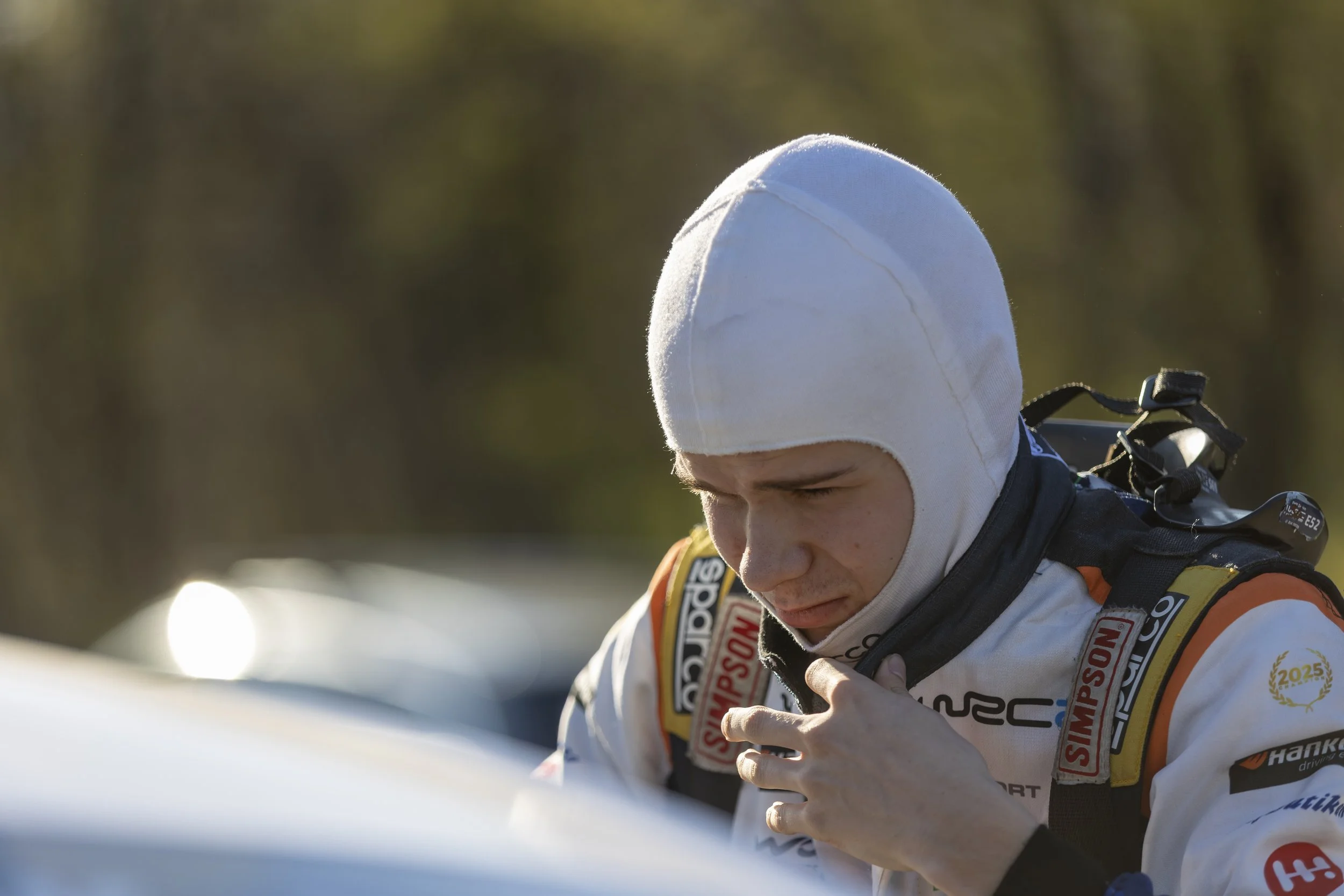Close-up of a race car driver adjusting his gear, wearing a white cap and a racing suit covered with sponsor patches.