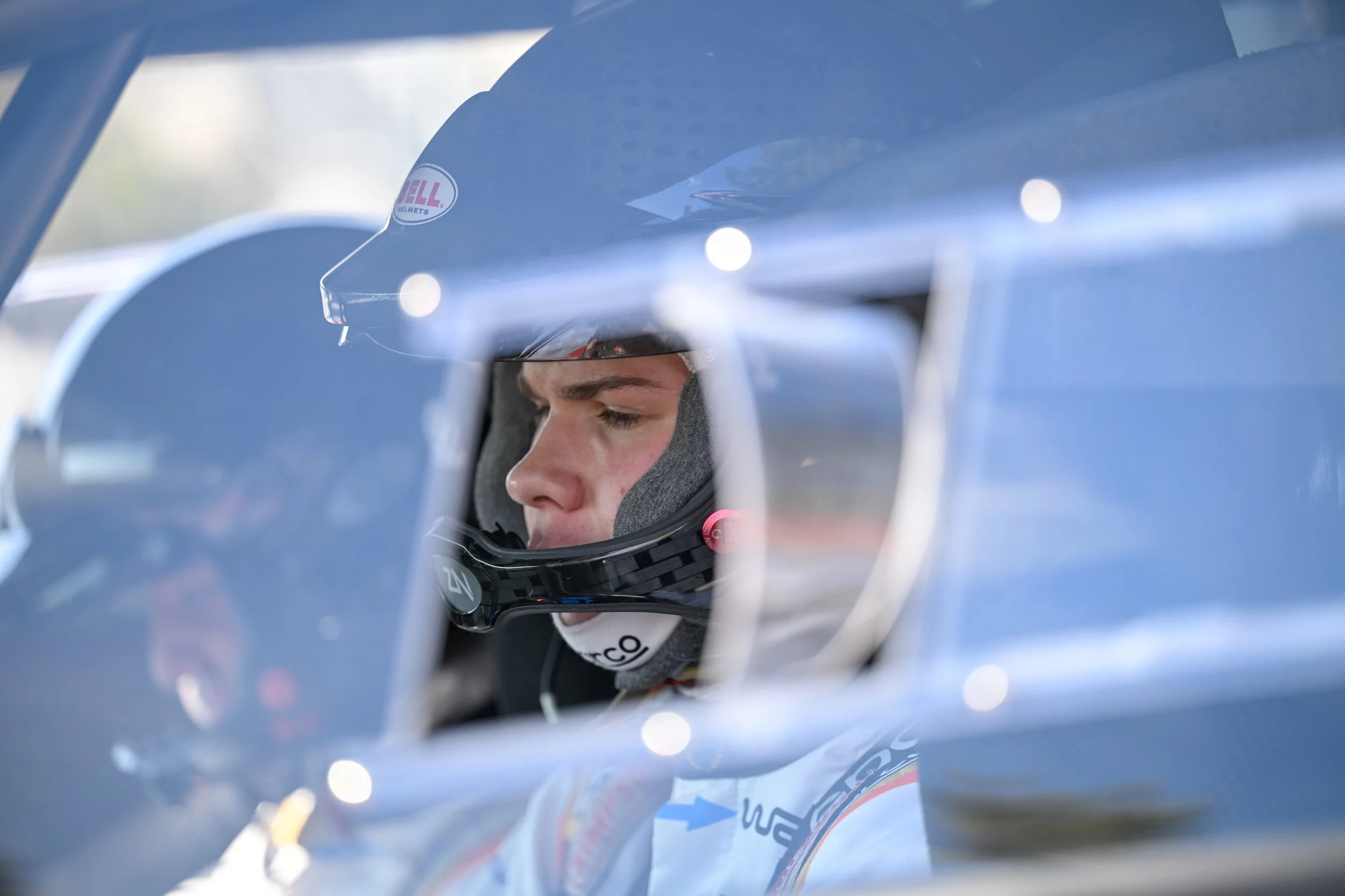 Close-up of a young male race car driver wearing a helmet, sitting in a race car.
