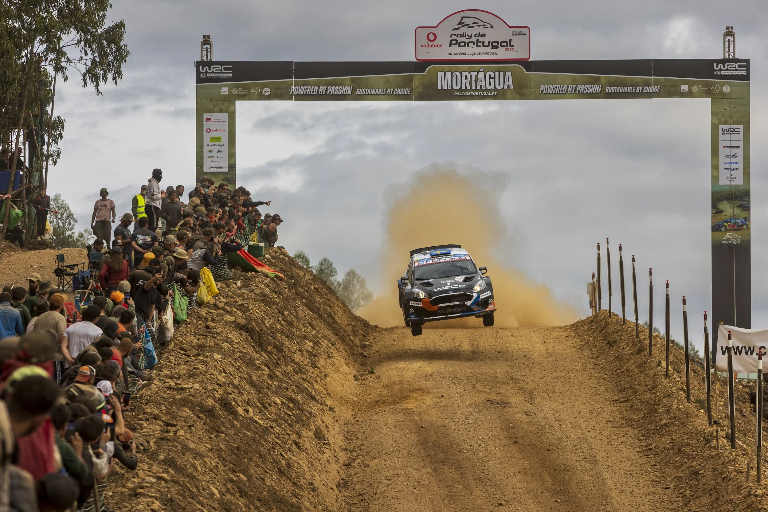 A rally car is airborne over a dirt track during a race, with spectators watching from a hillside and a large arch overhead that reads 'Rally de Portugal 2026'.