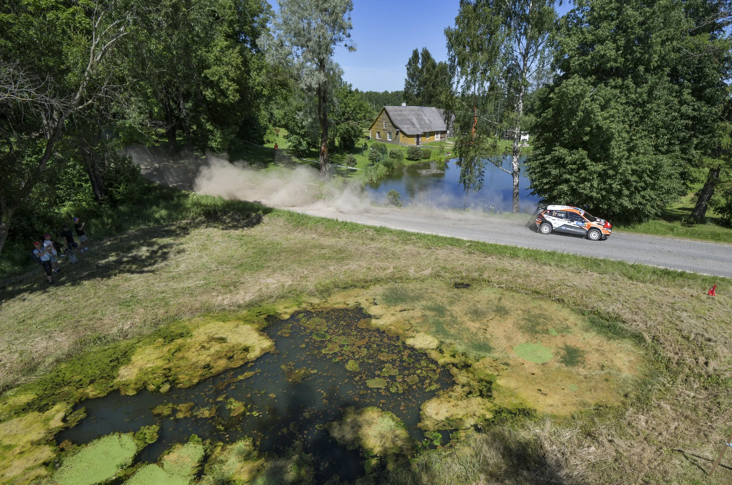 A rally car driving on a gravel road near a pond with green algae, in a green wooded area, with a house in the background and spectators on the left side watching.