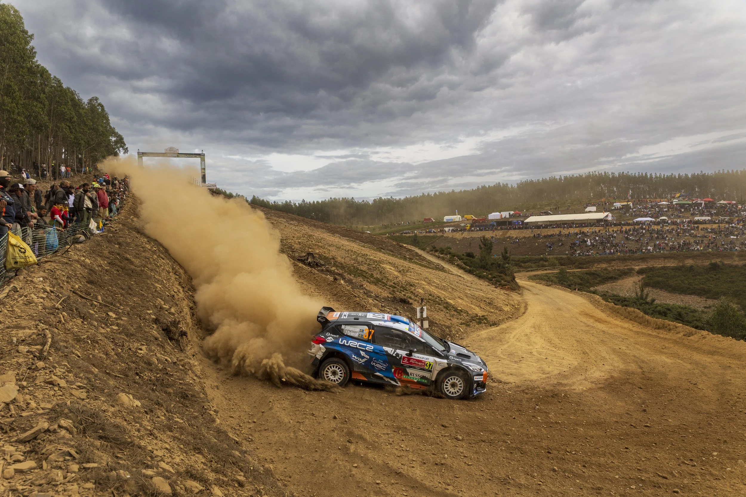 Rally cars racing on a dirt track, kicking up dust, with spectators watching from a hillside and the background showing a cloudy sky and distant packed stands.
