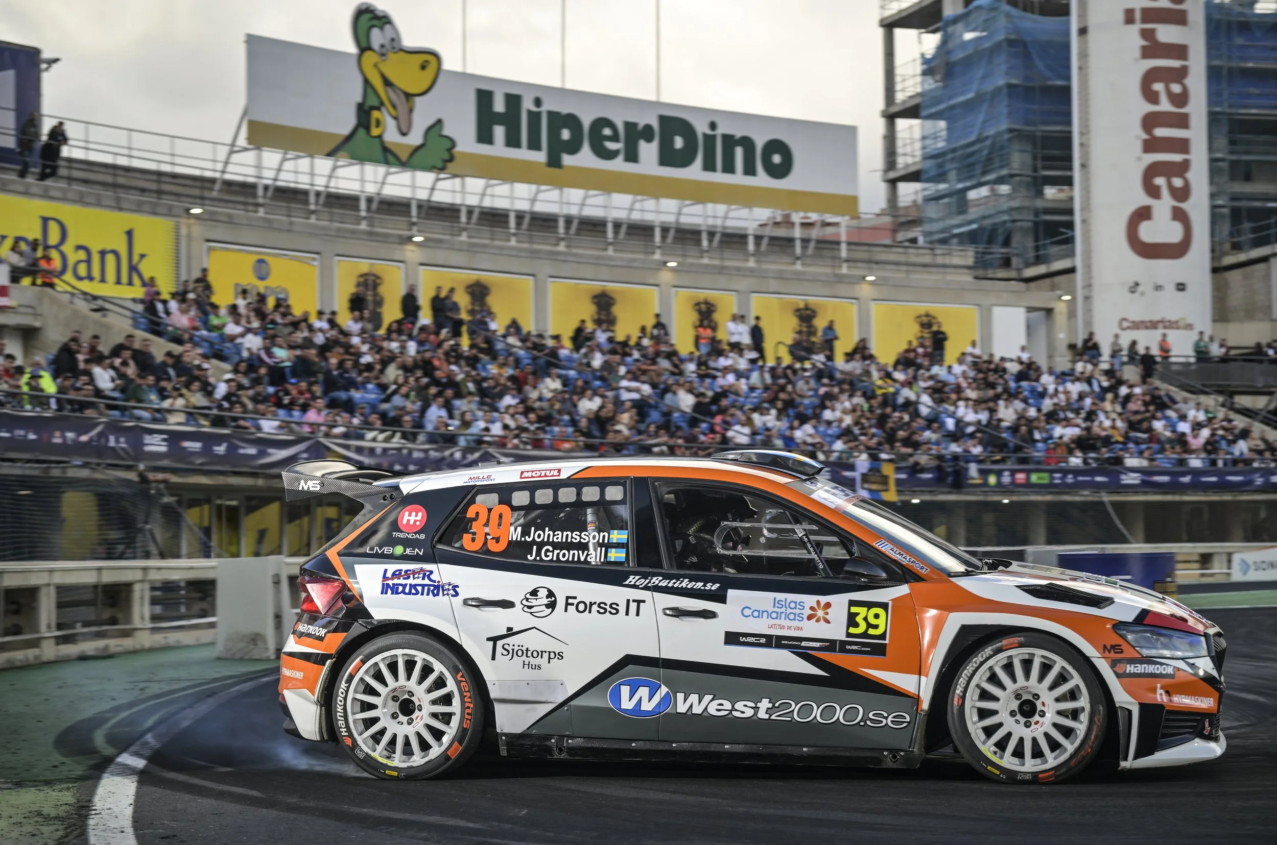 A rally car with the number 39 and Swedish flags on it, racing on a track with spectators in the background, advertising banners, and a large sign that reads "HiperDino".