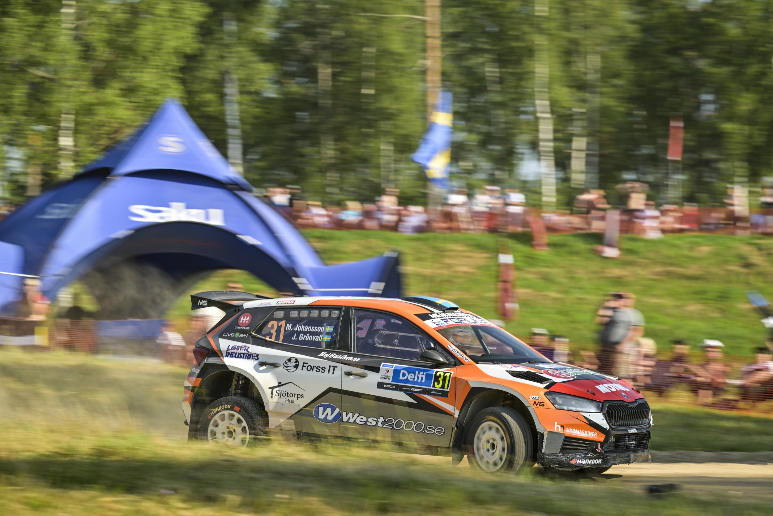 A rally car with the number 31 driven by M. Johansson and J. Grönvall speeding on a grassy track during a race, with a blurred background of spectators and flags.
