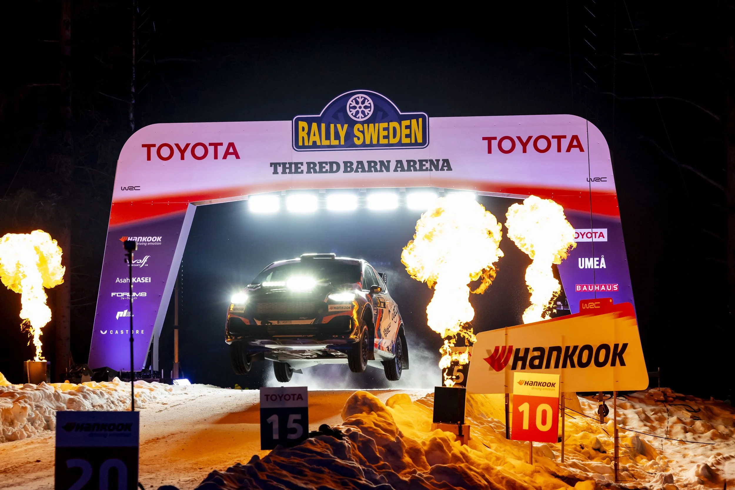 A rally car racing through snow at night during the Rally Sweden event, with flames and bright lights illuminating the scene.
