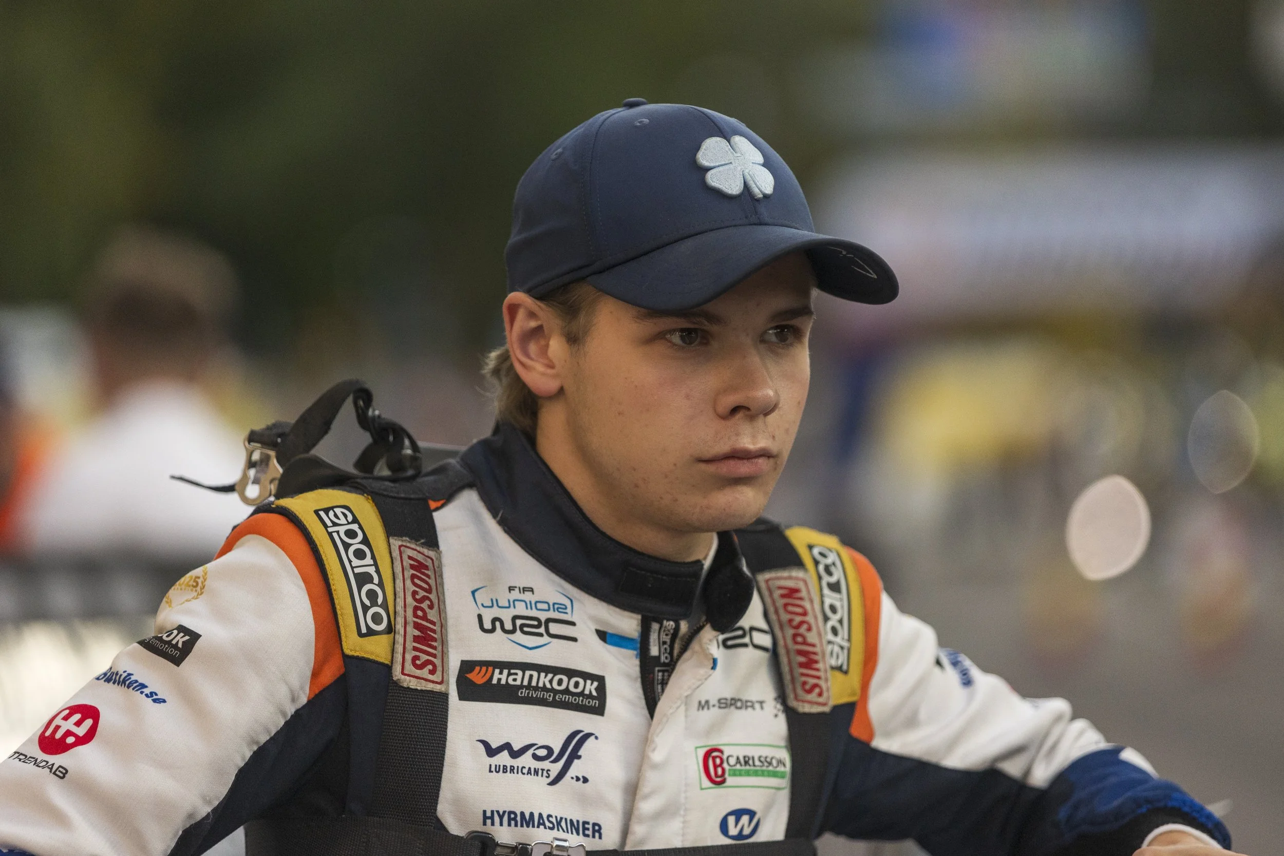 A race car driver wearing a white racing suit with sponsor patches and a dark blue cap with a white clover logo.