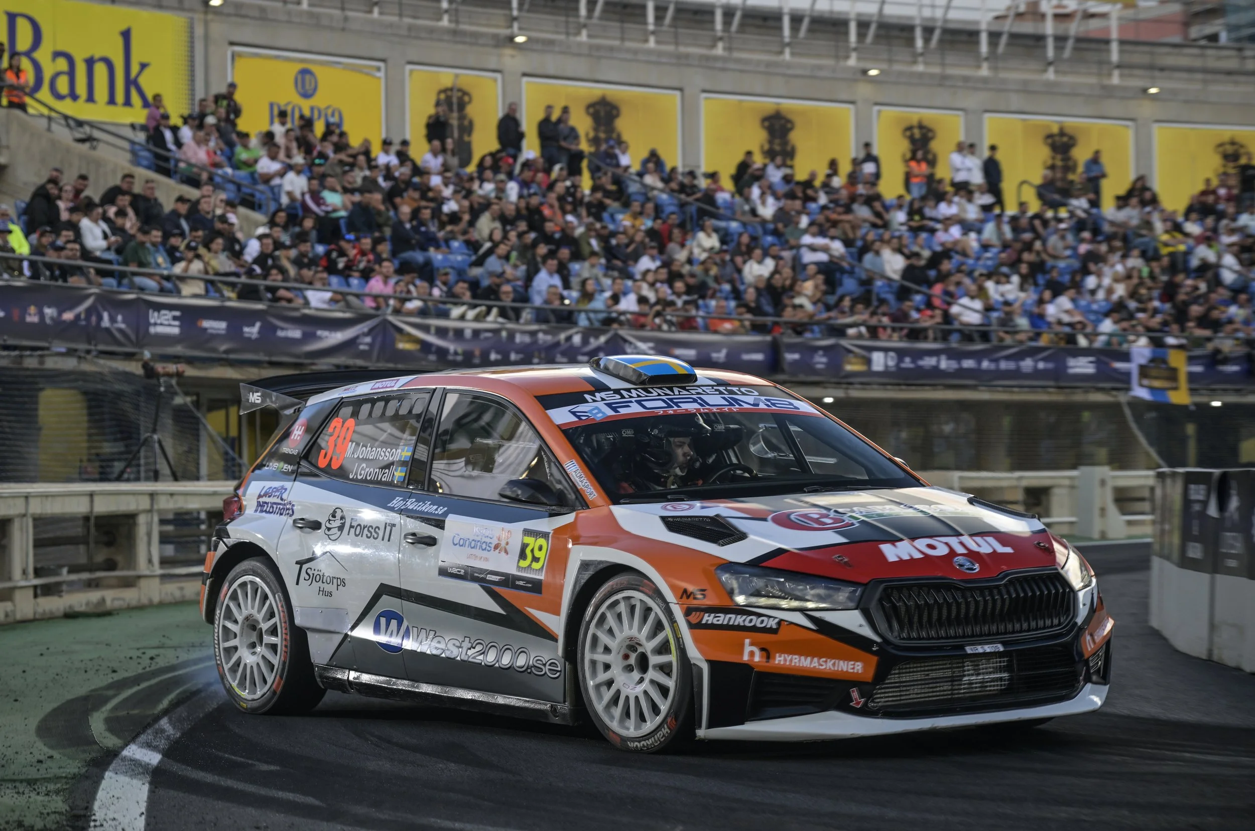 A rally car racing on a track with a crowd of spectators in the background.