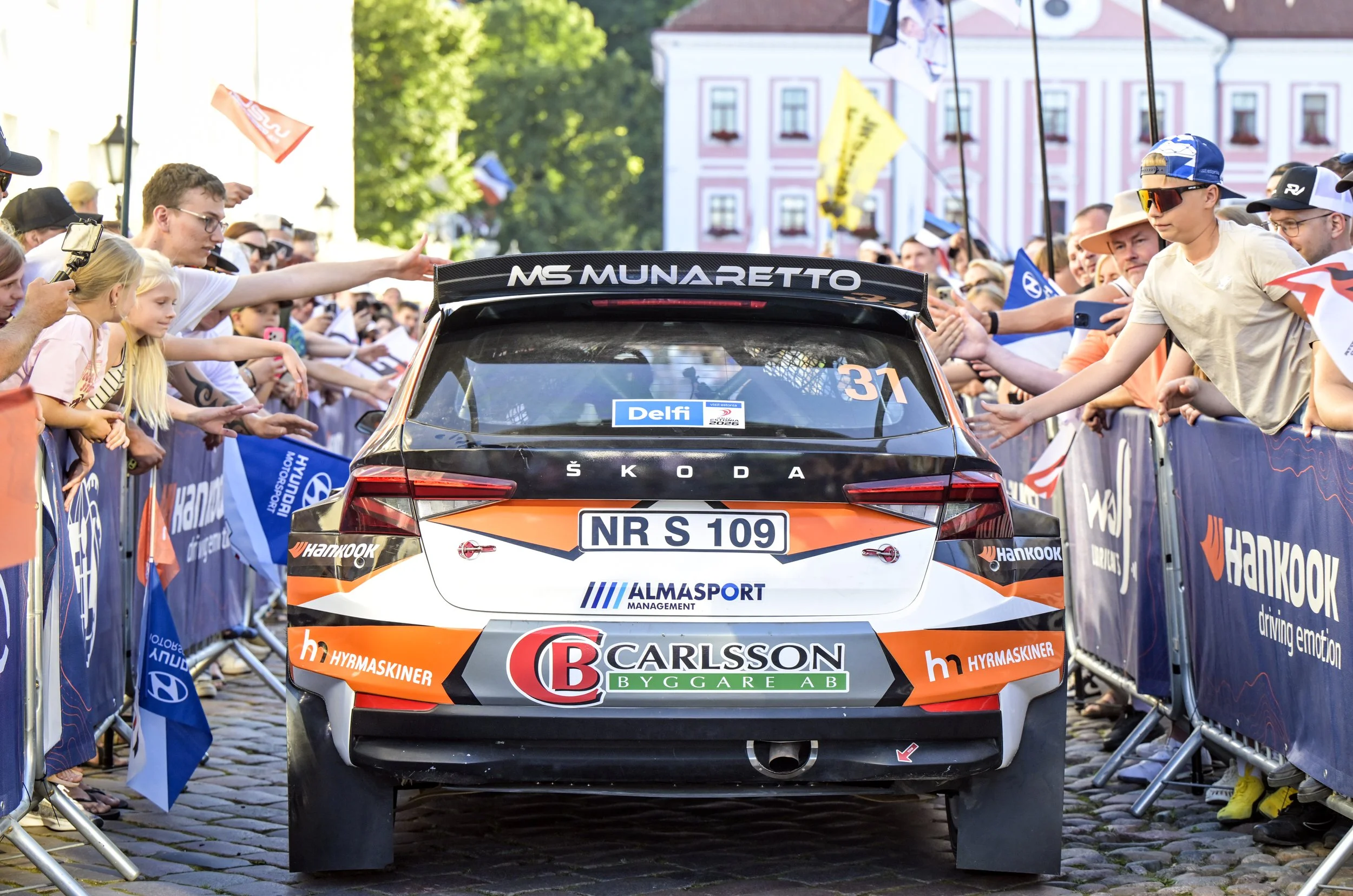 A black and orange rally car with sponsor logos and a large rear spoiler, stopped at a crowd of spectators behind barriers during a racing event. The crowd is reaching out towards the car, and flags are visible in the background.