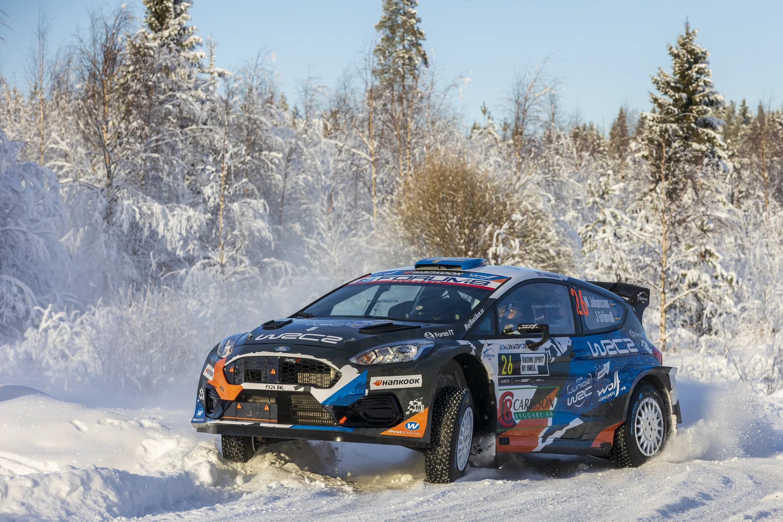 A black and blue rally car driving on a snowy winter landscape, surrounded by snow-covered trees and a clear blue sky.