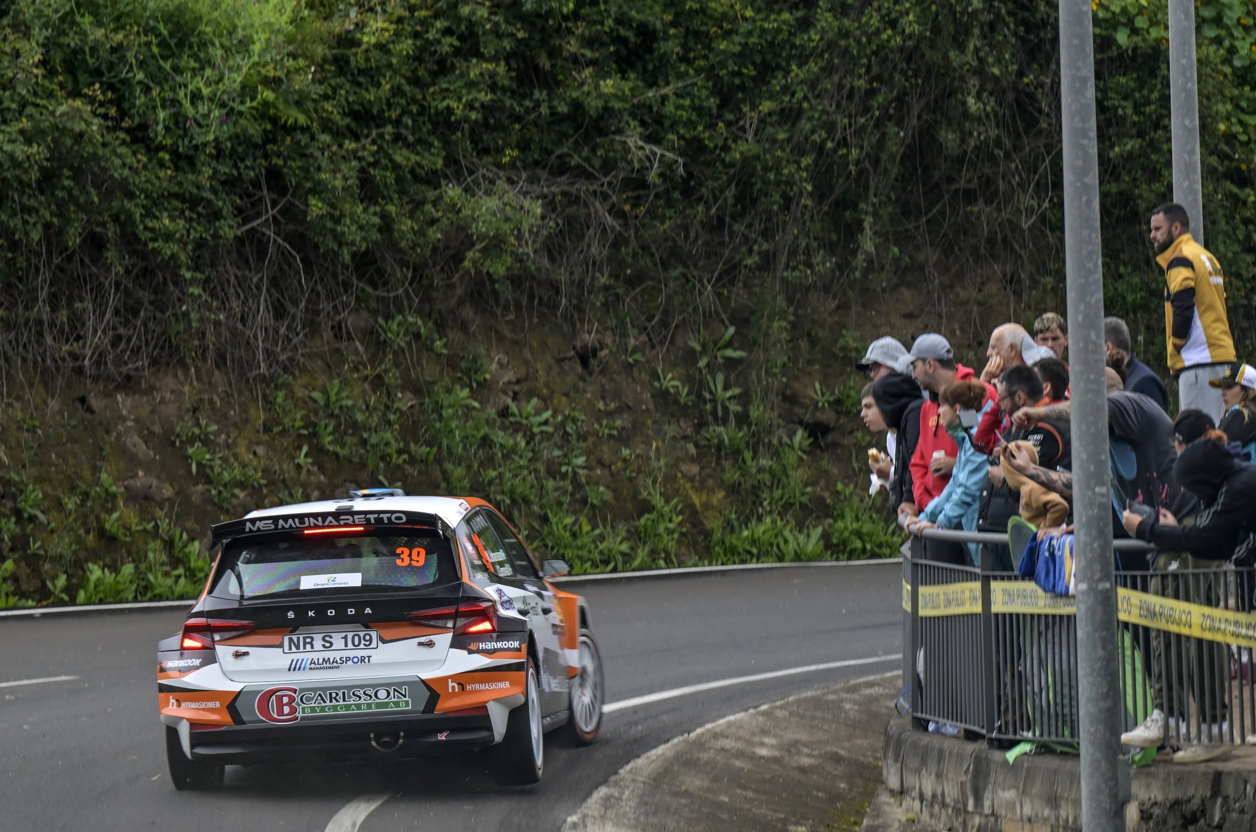 A rally car racing on a curved road with spectators behind a barrier watching from the side.