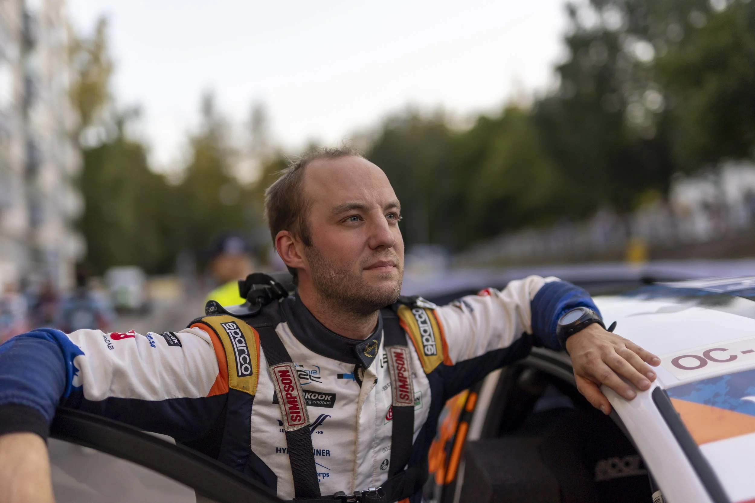 Motorsport driver wearing a racing suit and harness, sitting in a race car with arms outstretched, in an outdoor setting with trees and blurred background.