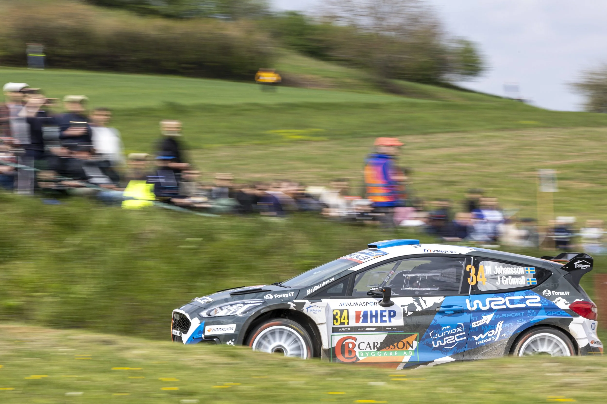 A rally car race with a black and blue car in the foreground, displaying the number 34 and sponsorship logos, speeding along a grassy course with a blurred audience and landscape in the background.