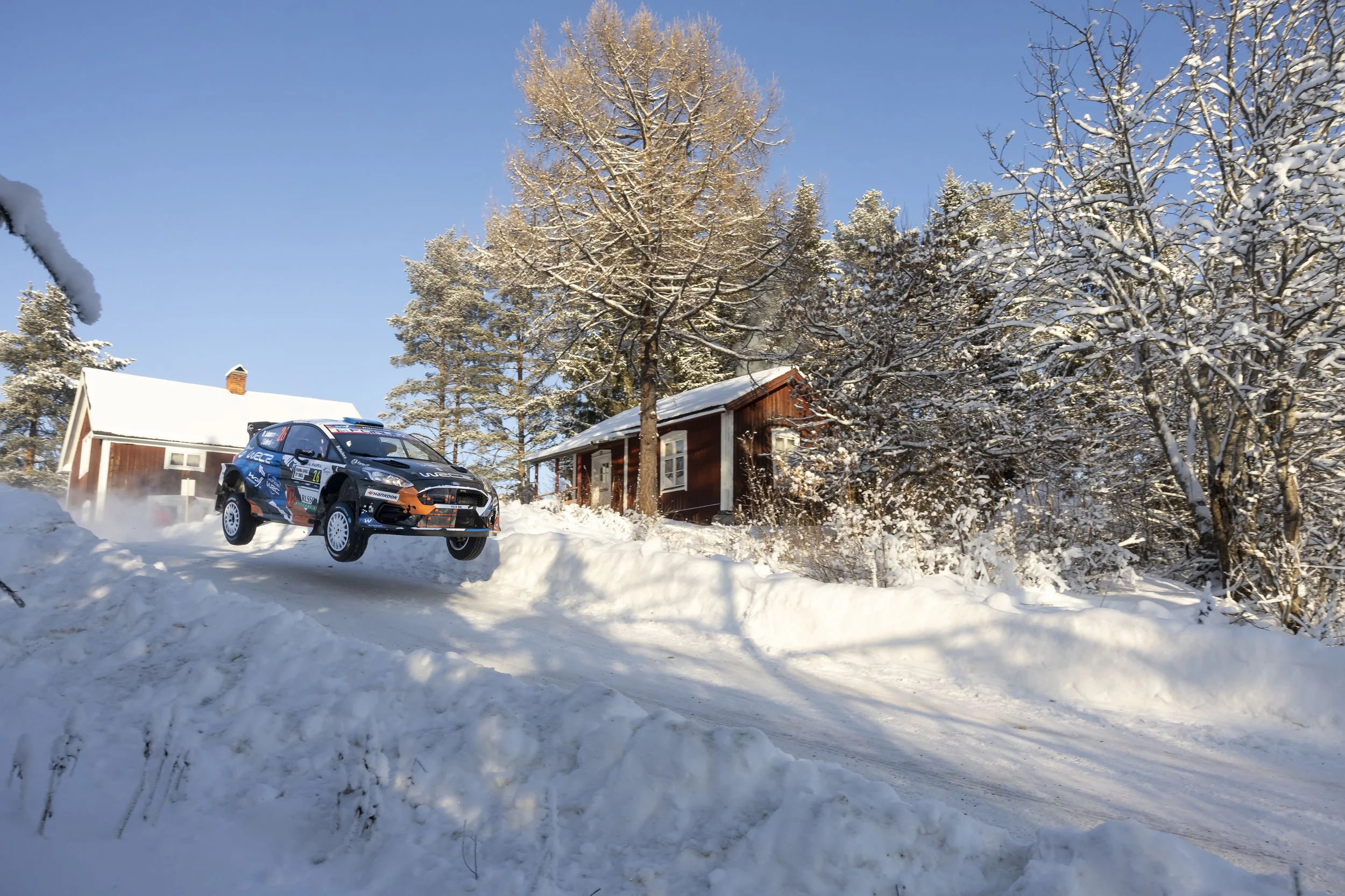 A snowy landscape with a rally car driving quickly through the snow, kicking up snow behind it. There are snow-covered trees and a wooden house in the background, under a clear blue sky.