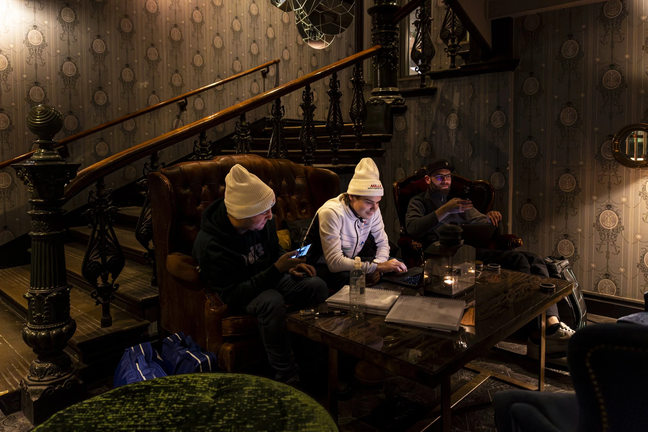 Four people sitting on a vintage leather sofa at a table in a dimly lit room with ornate wallpaper, a staircase, and a decorative light fixture overhead. Three men are using electronic devices, with water bottles and notepads on the table.