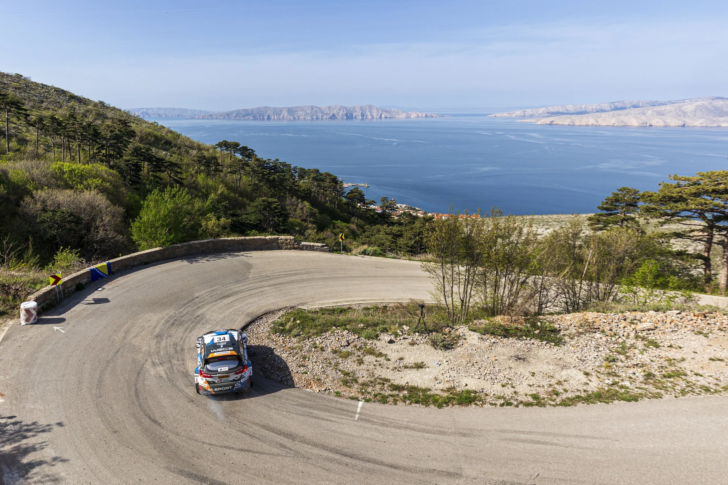 Race cars on a winding mountain road overlooking a large body of water and distant hills or mountains.