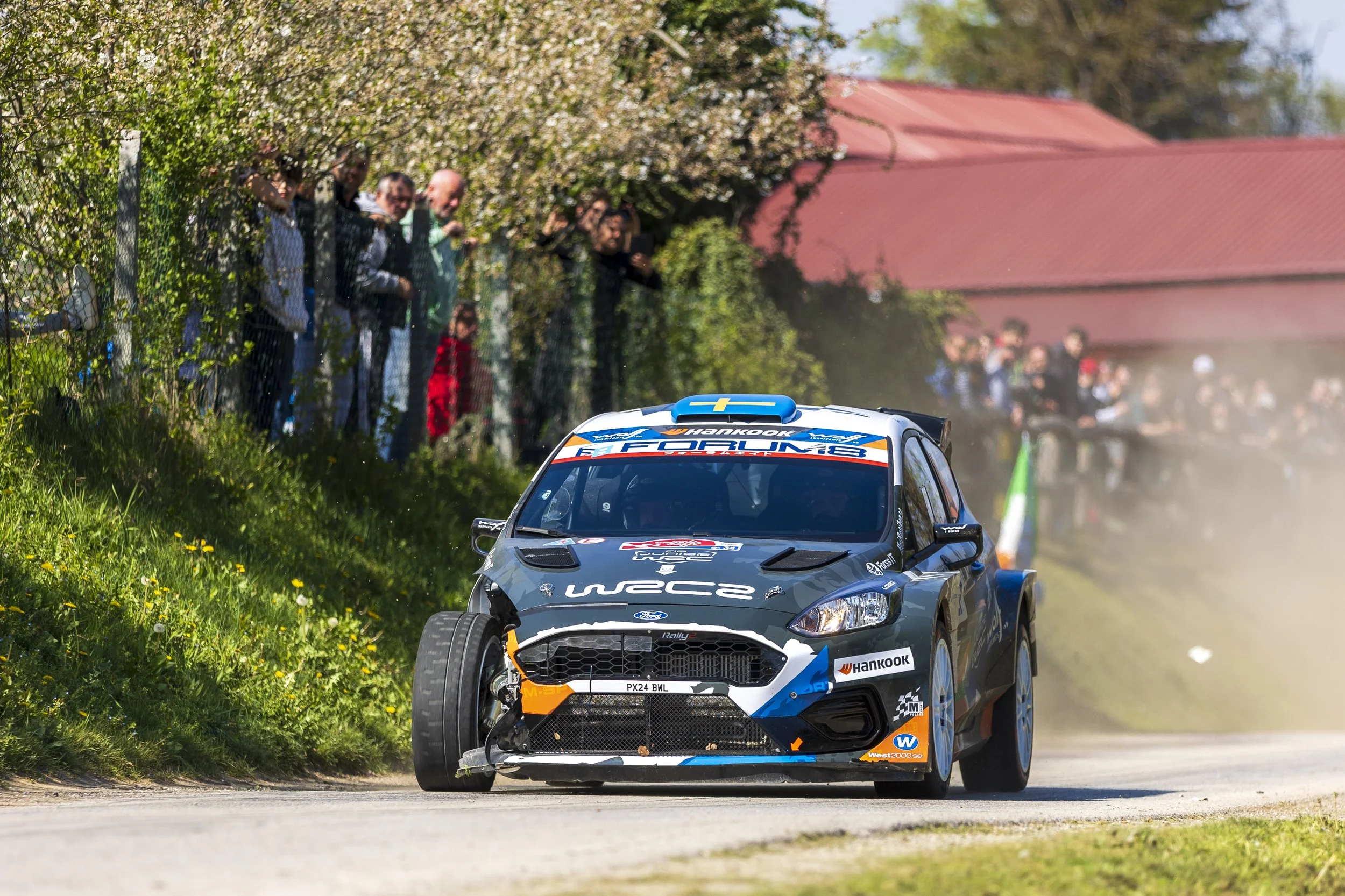 A rally car speeds down a dirt road, kicking up dust, with a crowd of spectators watching from behind a fence under flowering trees and a red-roofed building in the background.