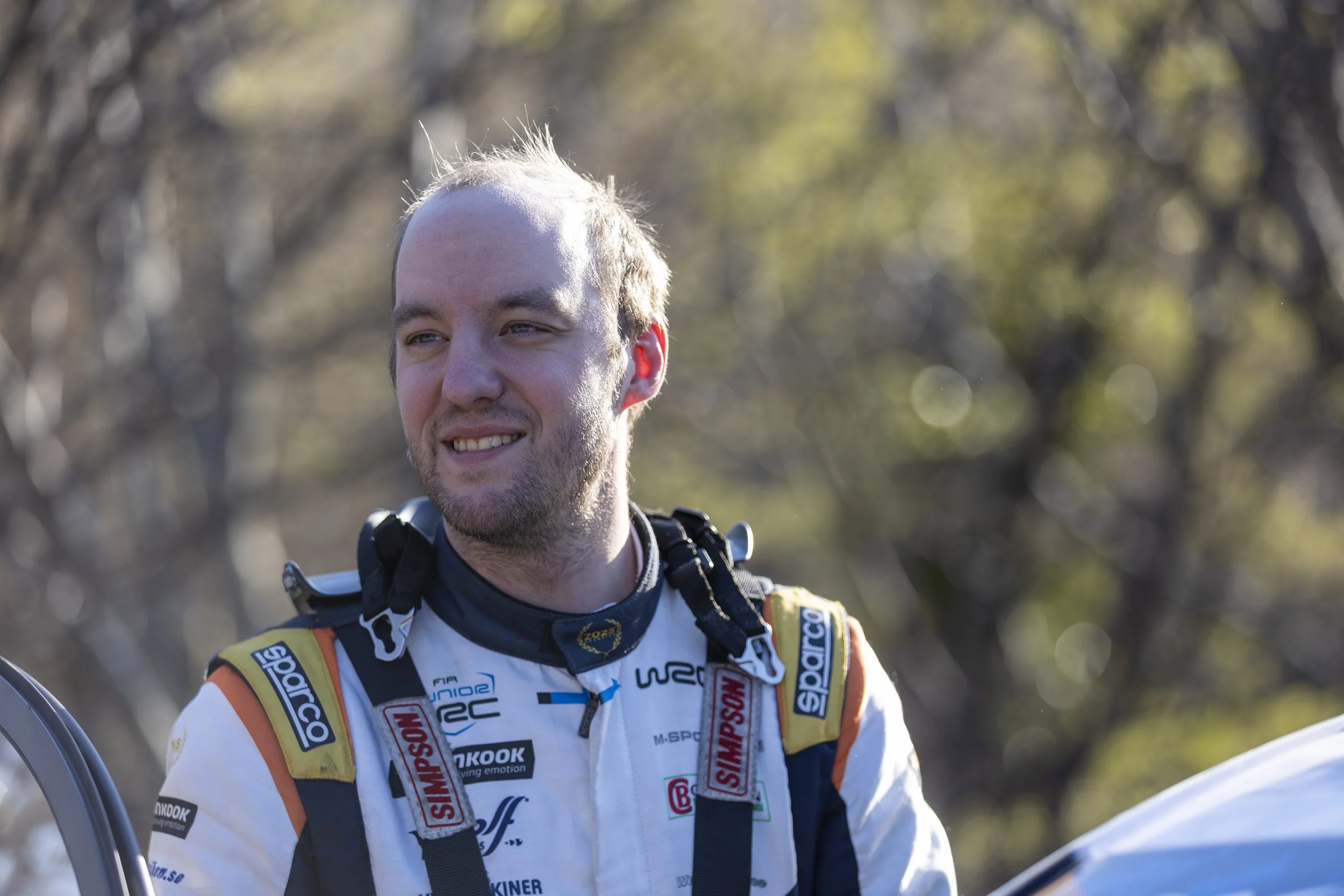 A man dressed in race gear with sponsor logos, smiling outdoors with trees in the background.