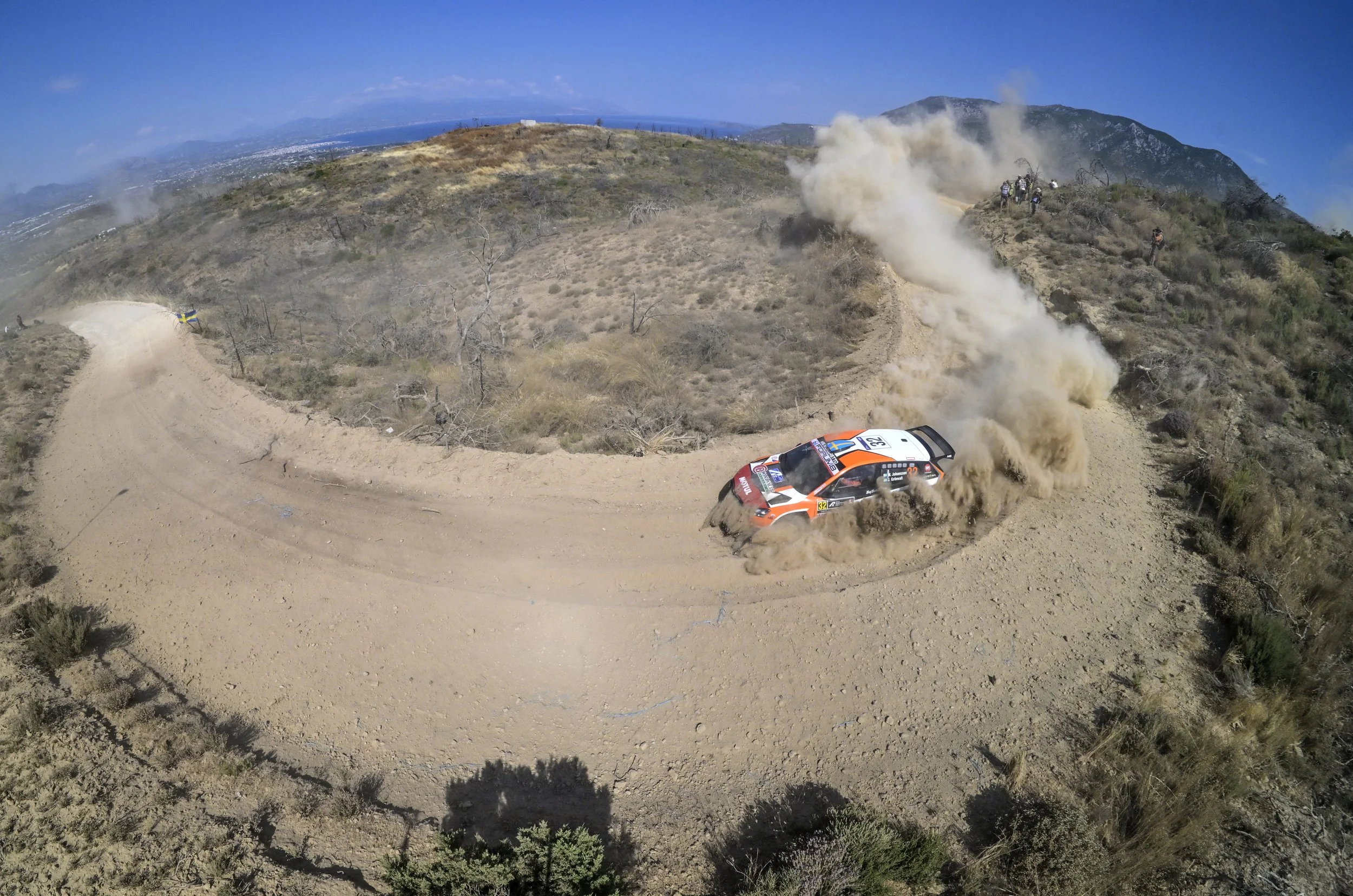 A rally car kicking up dust and dirt along a dirt trail on a mountain ridge, with a distant landscape and blue sky in the background.