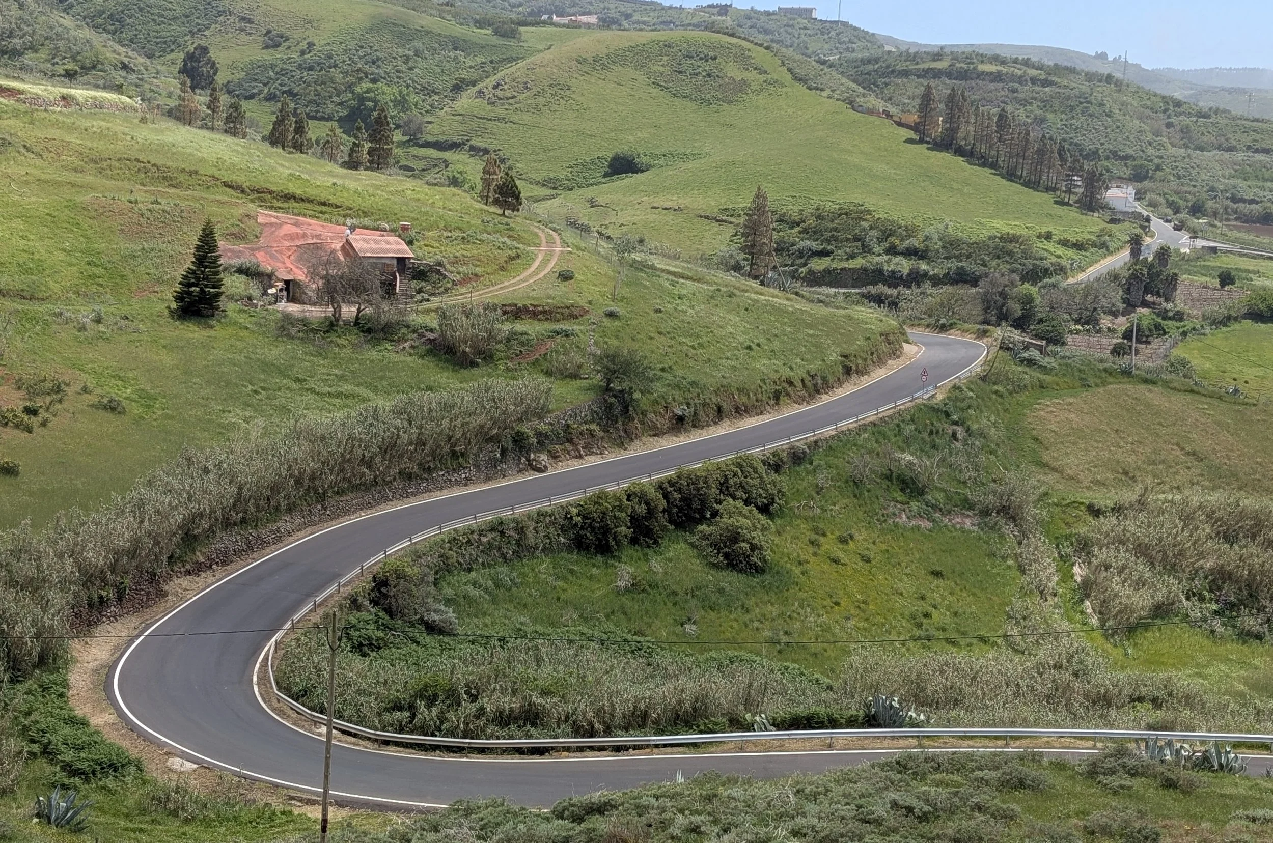 A winding paved road moving through green hills with trees, shrubs, and small houses scattered across the landscape, under a clear blue sky.