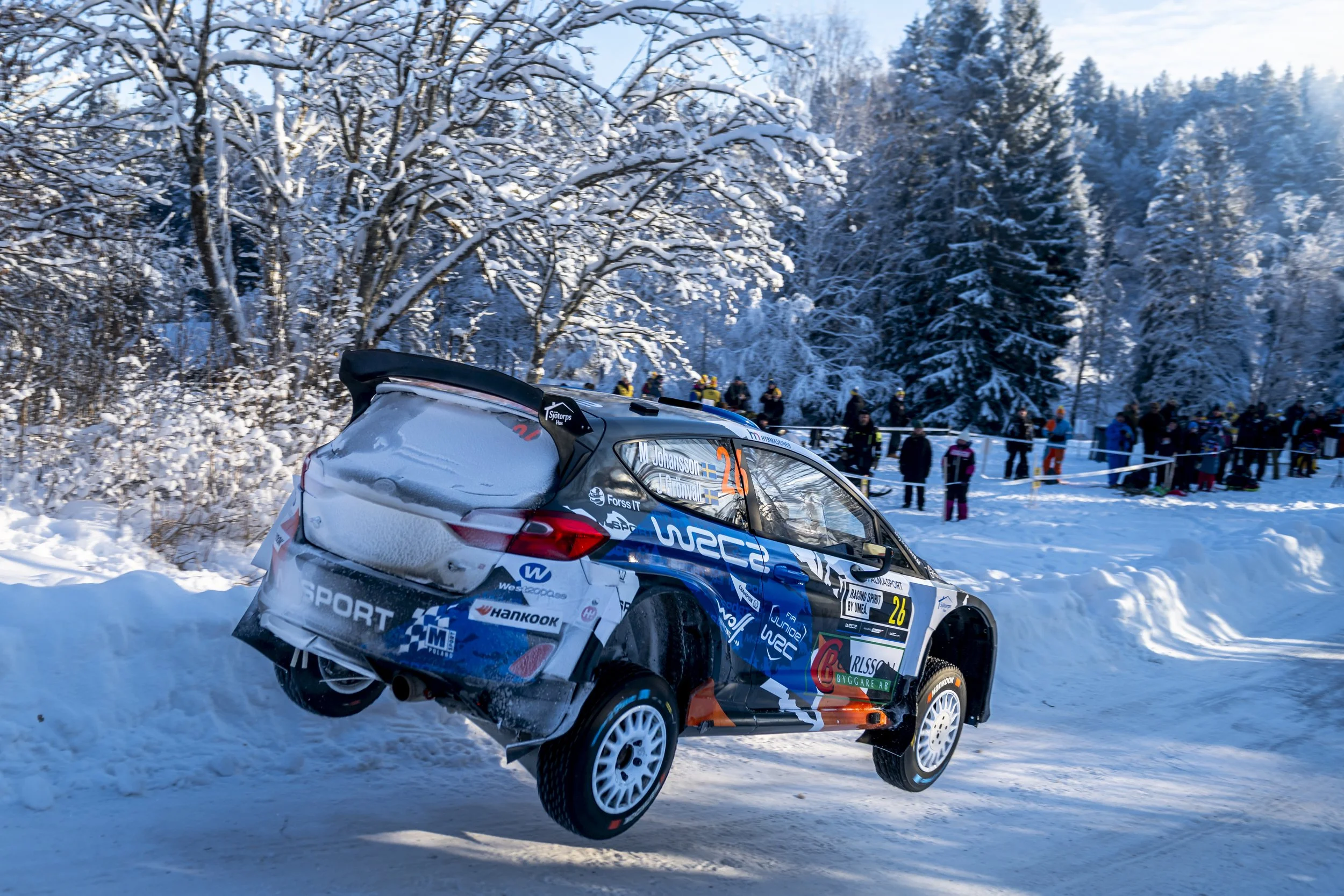 A rally car in mid-air during a winter race, with snow-covered trees in the background and spectators watching from the side.