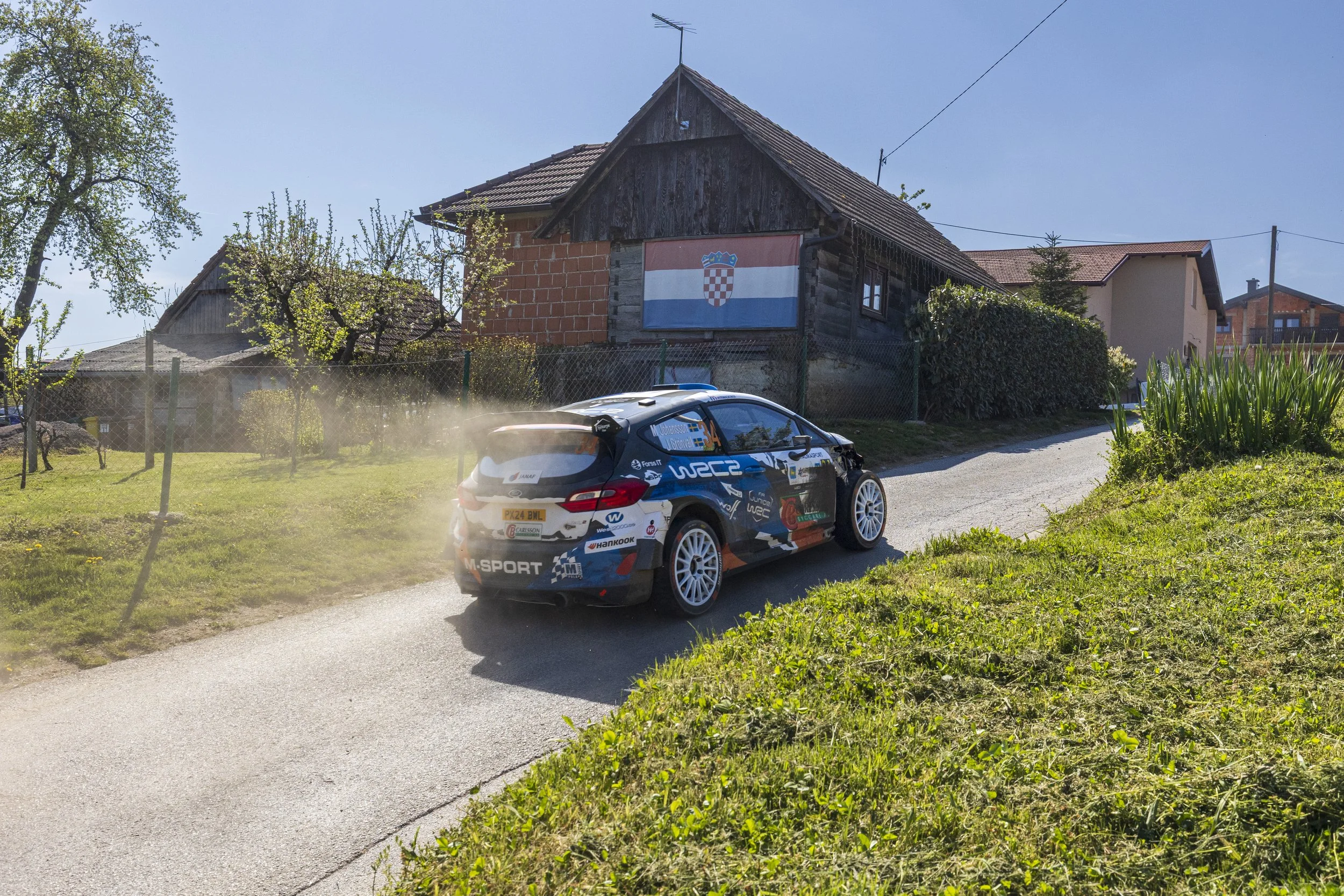 A rally car speeding on a dirt road beside a grassy yard with trees and houses, with a Croatian flag hanging on a house in the background.