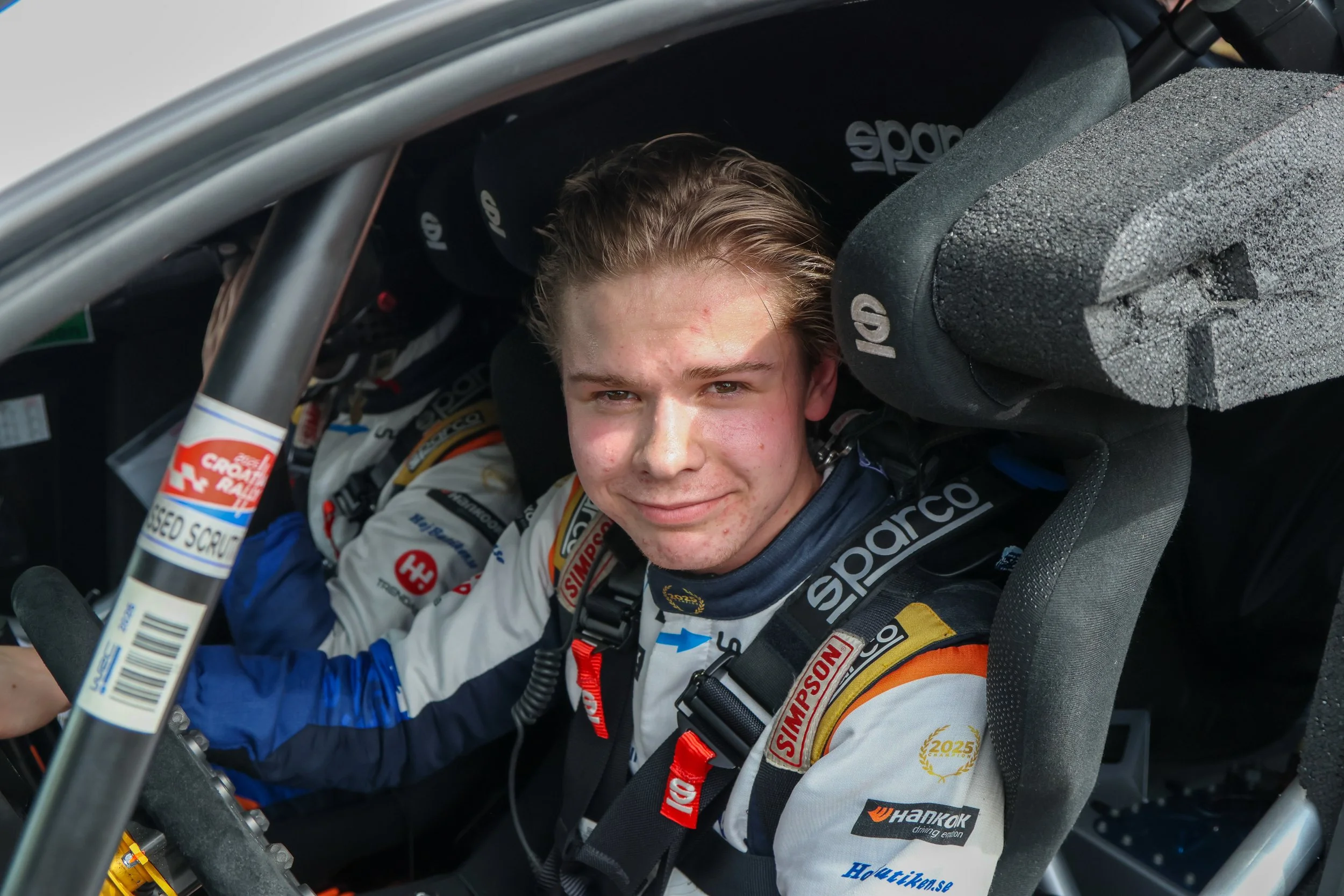 A young race car driver with blonde hair sitting inside a race car, smiling. He is wearing a racing suit with sponsor patches and a black safety harness. The car's interior and window frame are visible.