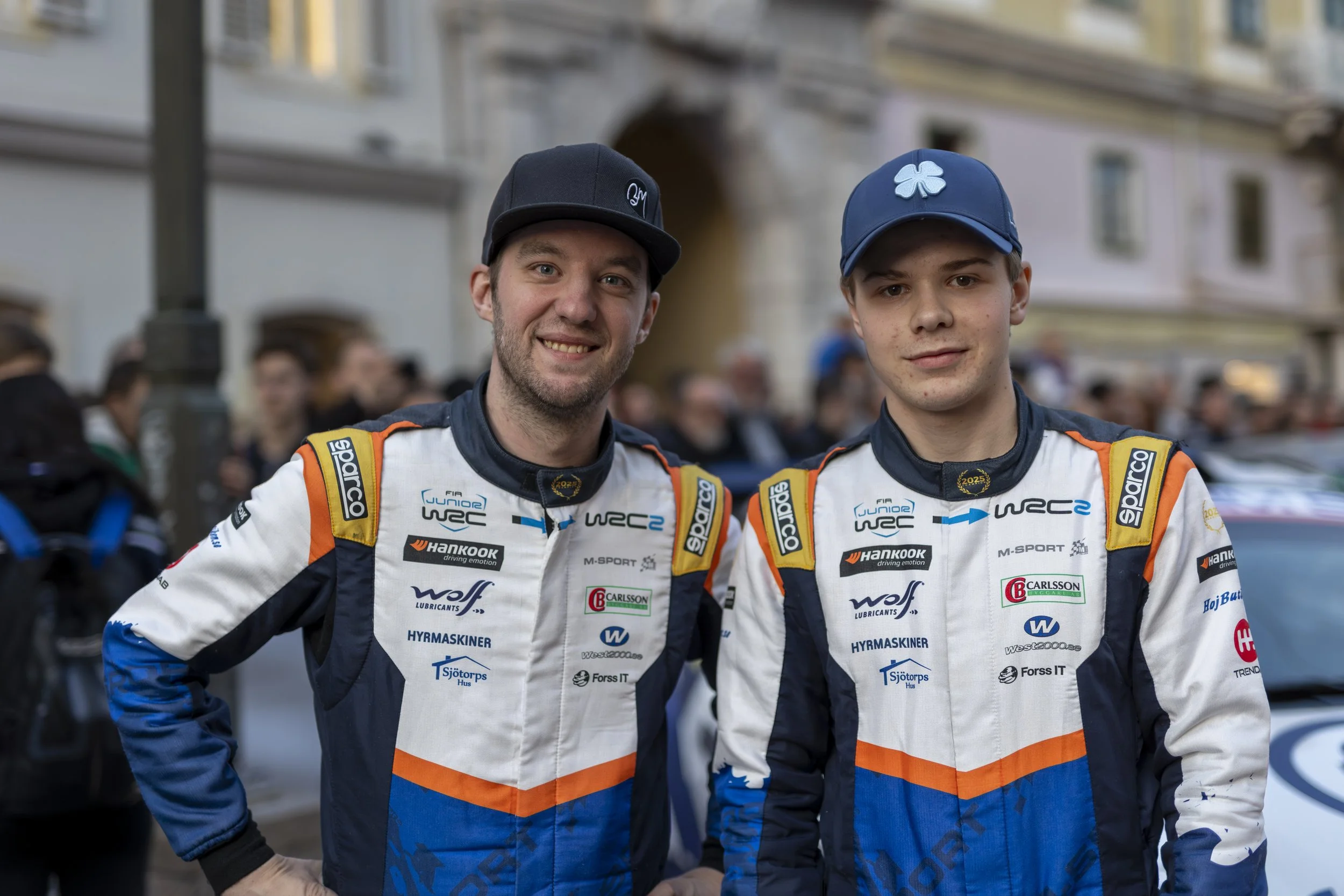 Two young male race car drivers in white and blue racing suits with sponsor logos, standing close together with their arms around each other, smiling at the camera during daytime at a public outdoor event.