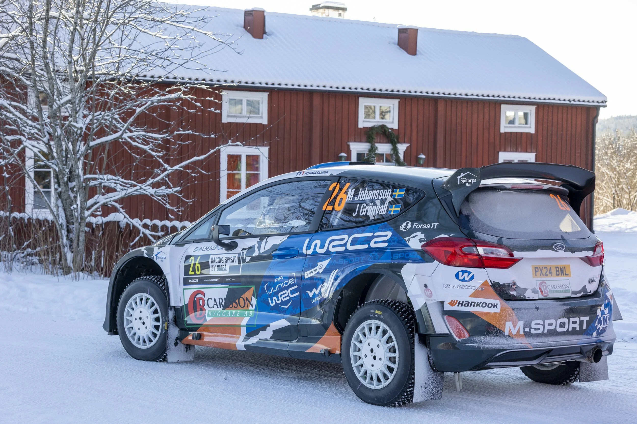 A rally car covered in sponsor logos parked on a snow-covered ground with a red wooden house and a snow-covered tree in the background.