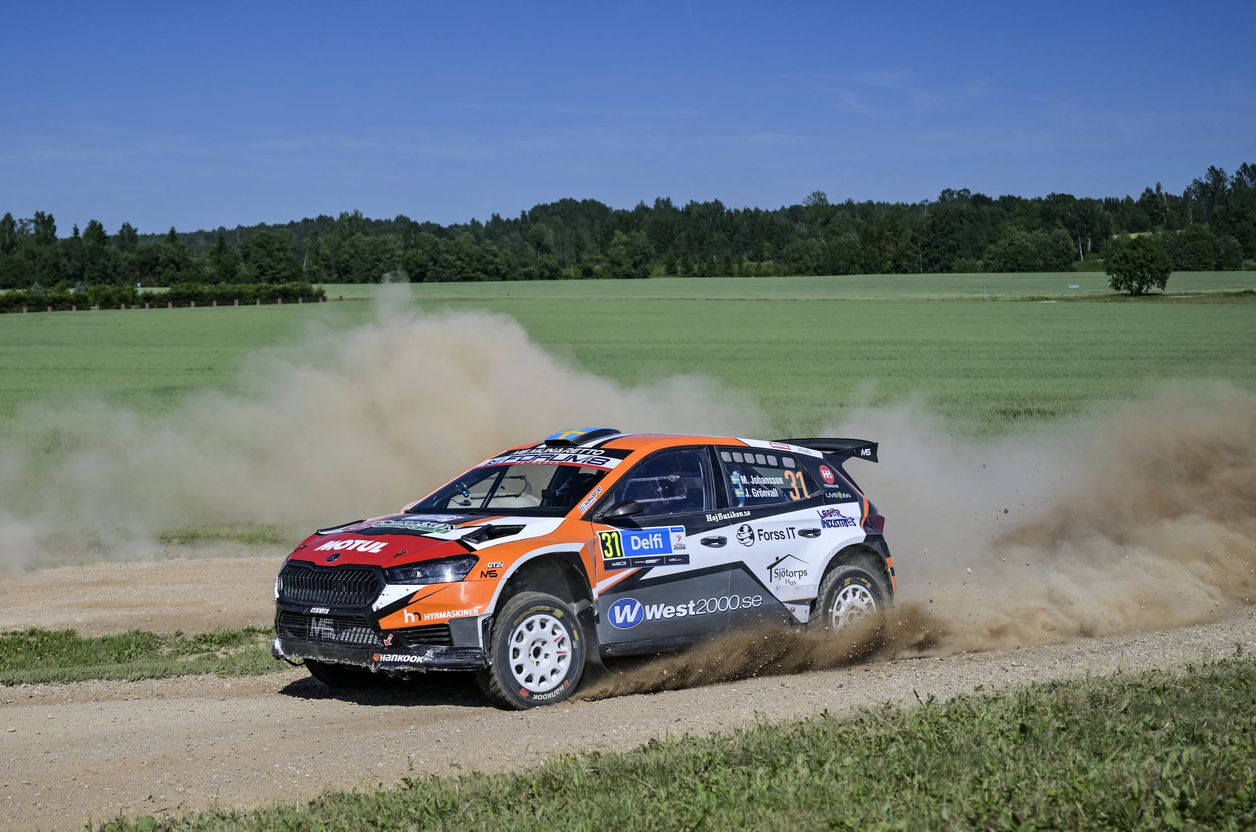 A rally car racing on a dirt track with dust clouds, surrounded by green grass and trees under a blue sky.