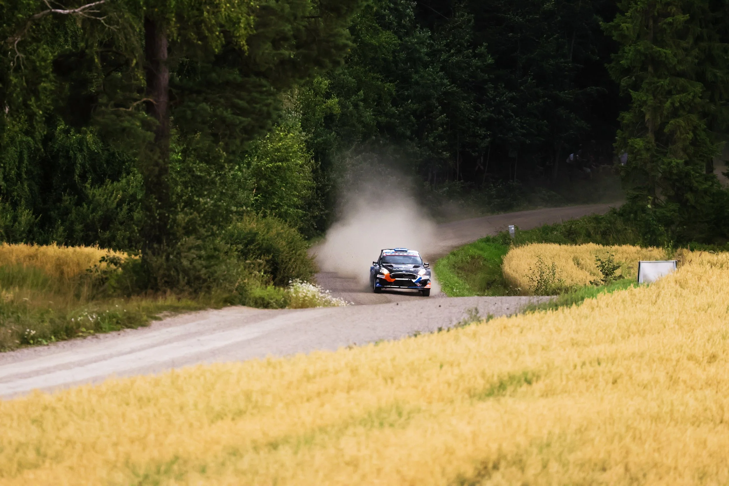A rally car driving on a dirt road through a forest with clouds of dust behind it.