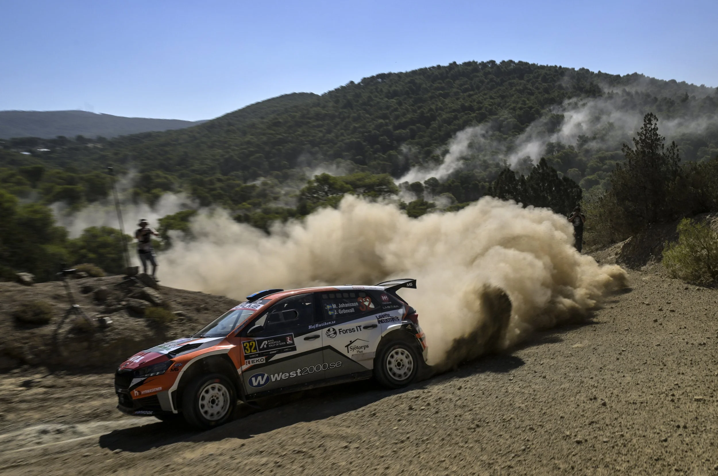 A rally car racing on a dirt mountain road, kicking up dust with a green forested hillside in the background under a clear blue sky.