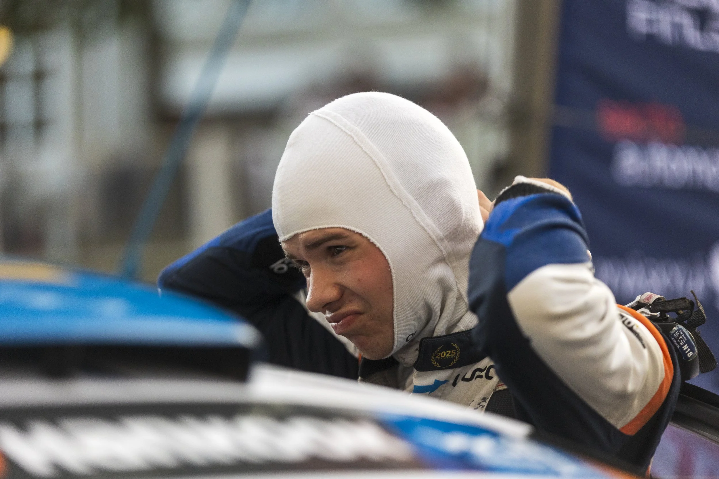 A smiling race car driver in a white racing suit and white helmet leans over a race car, with a blurred background of a pit area.