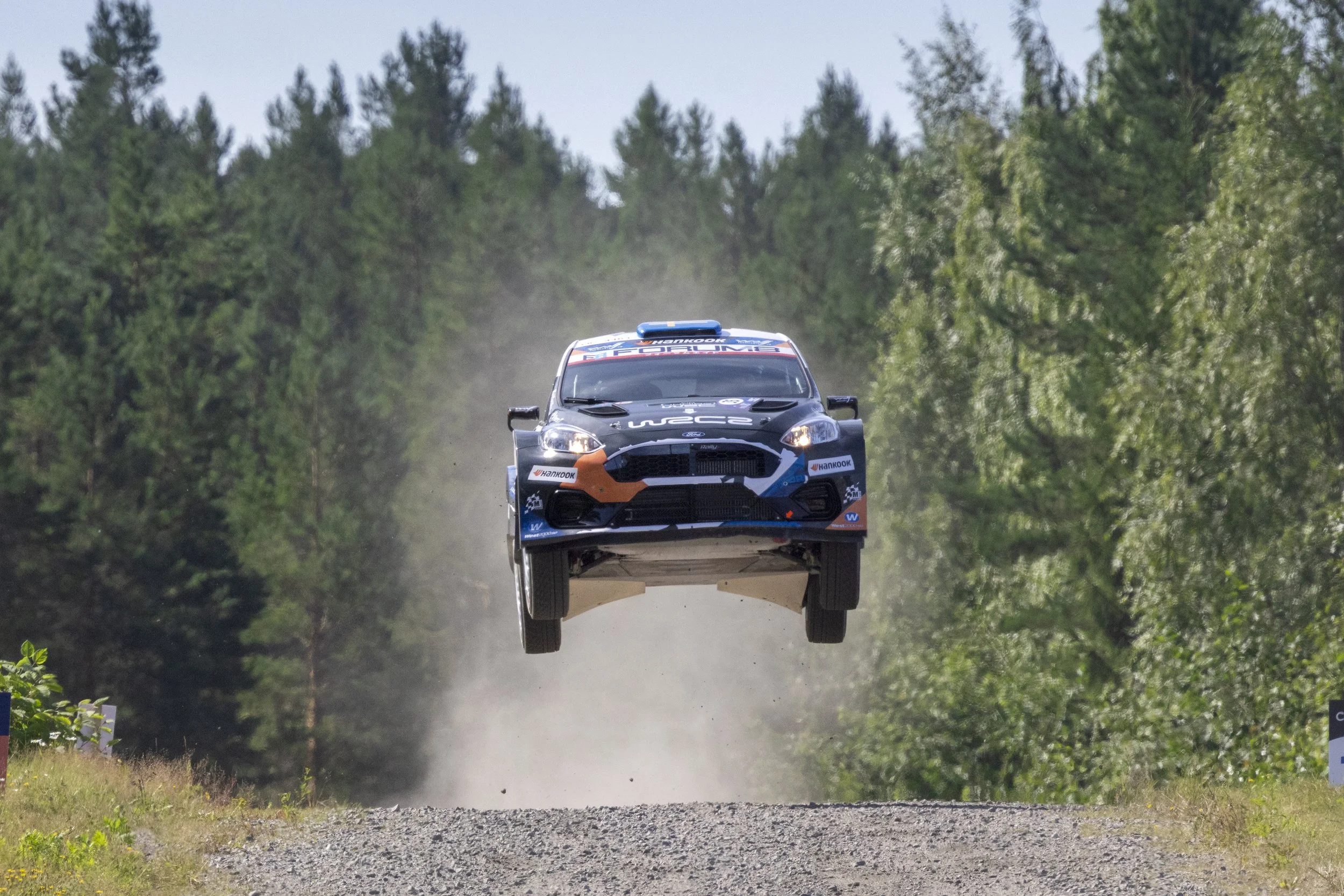 A rally car in mid-air after jumping off a dirt track surrounded by trees.