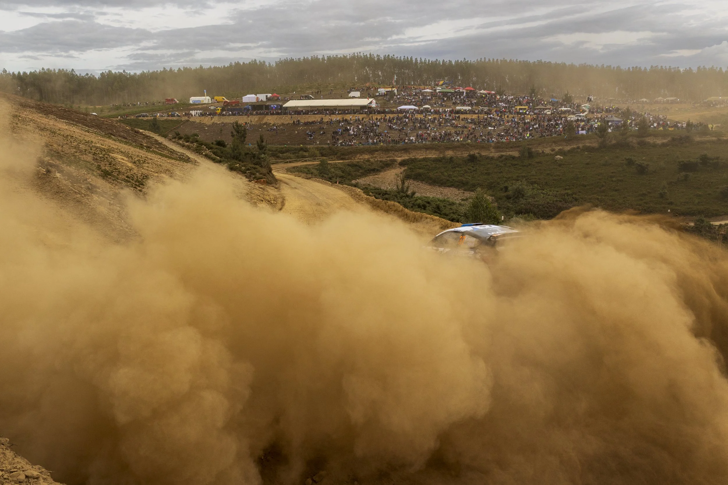 A car racing on a dirt track during a rally, with a large crowd of spectators and tents on a hillside in the background, dust clouds behind the car indicating high speed.