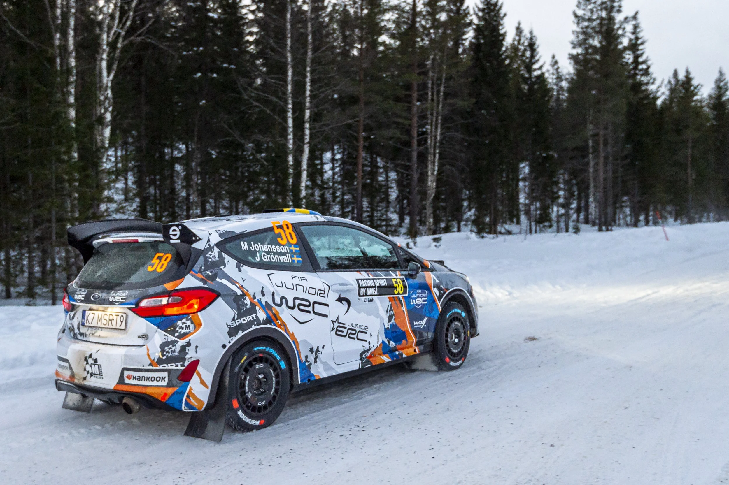 A rally car driving on a snowy road near a forest, with snow-covered ground and tall pine trees in the background.