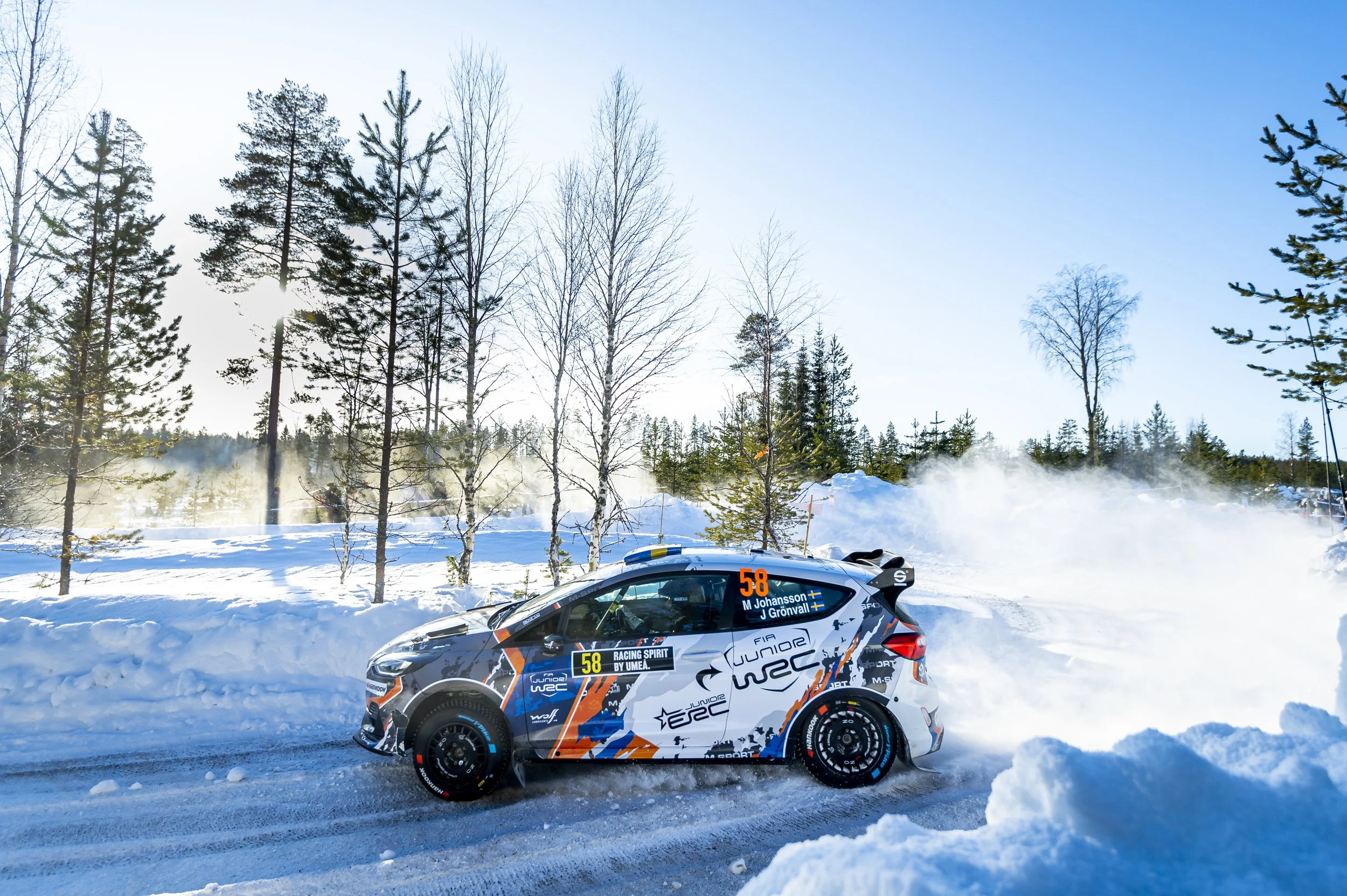 A rally car with racing decals and the number 58 on the side, driving on a snow-covered road with trees in the background. The sky is clear and sunlight shines through the trees.