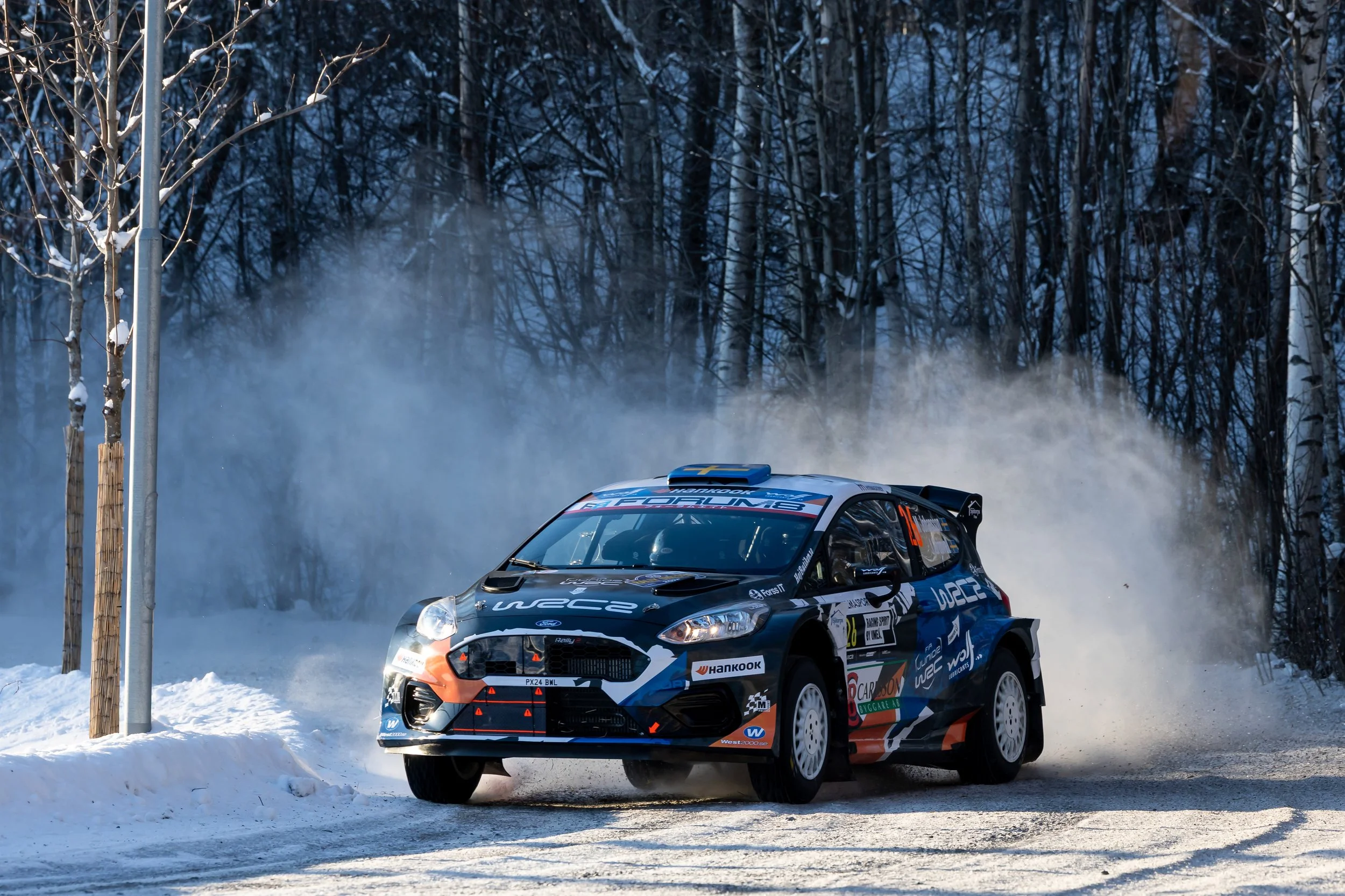 A rally car speeding on a snowy road, kicking up snow and mist, with a dense forest in the background during winter.