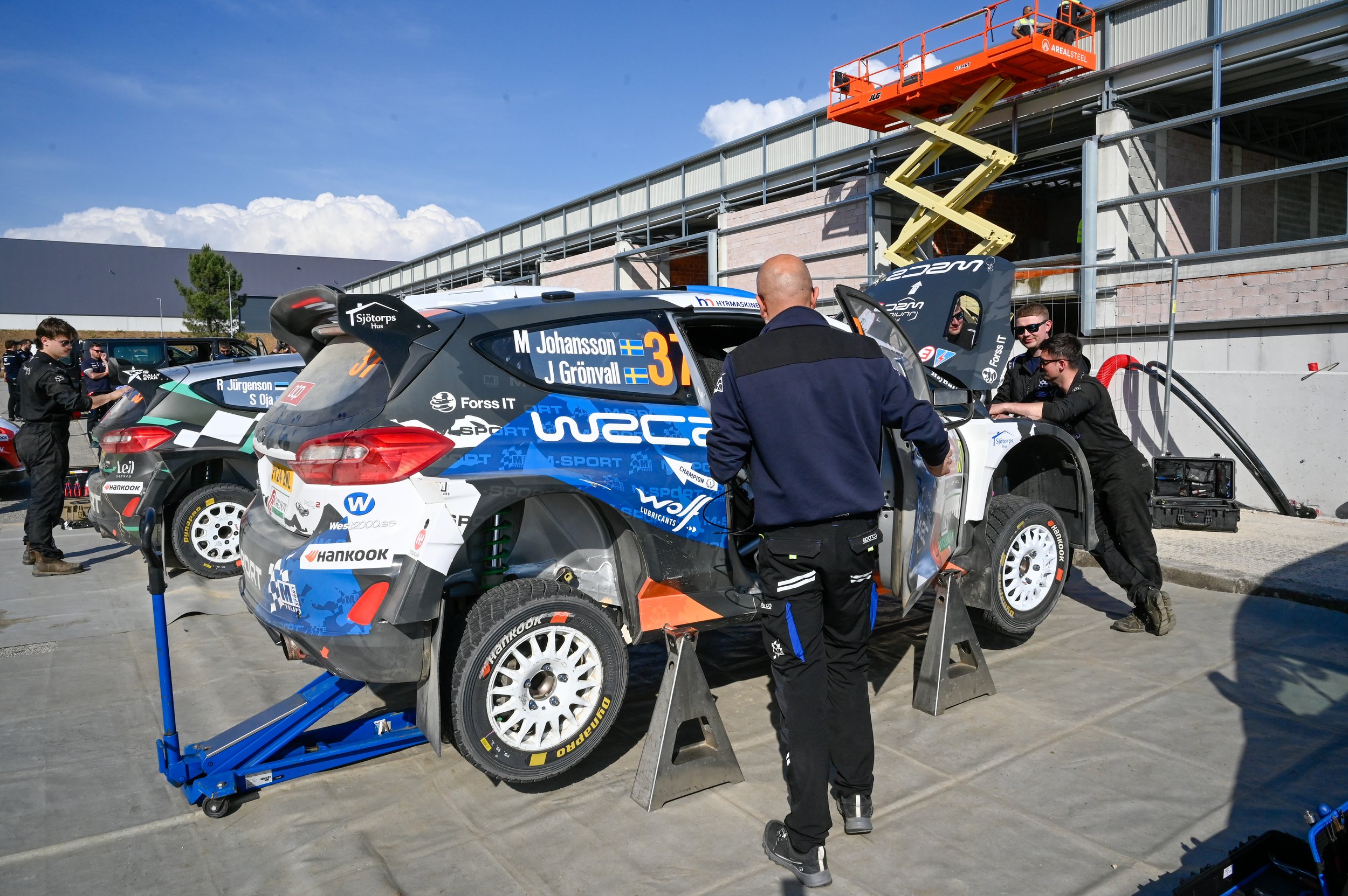 Race cars in a pit stop area being serviced by mechanics during a race event, with a building under construction and a scissor lift in the background under a clear blue sky.