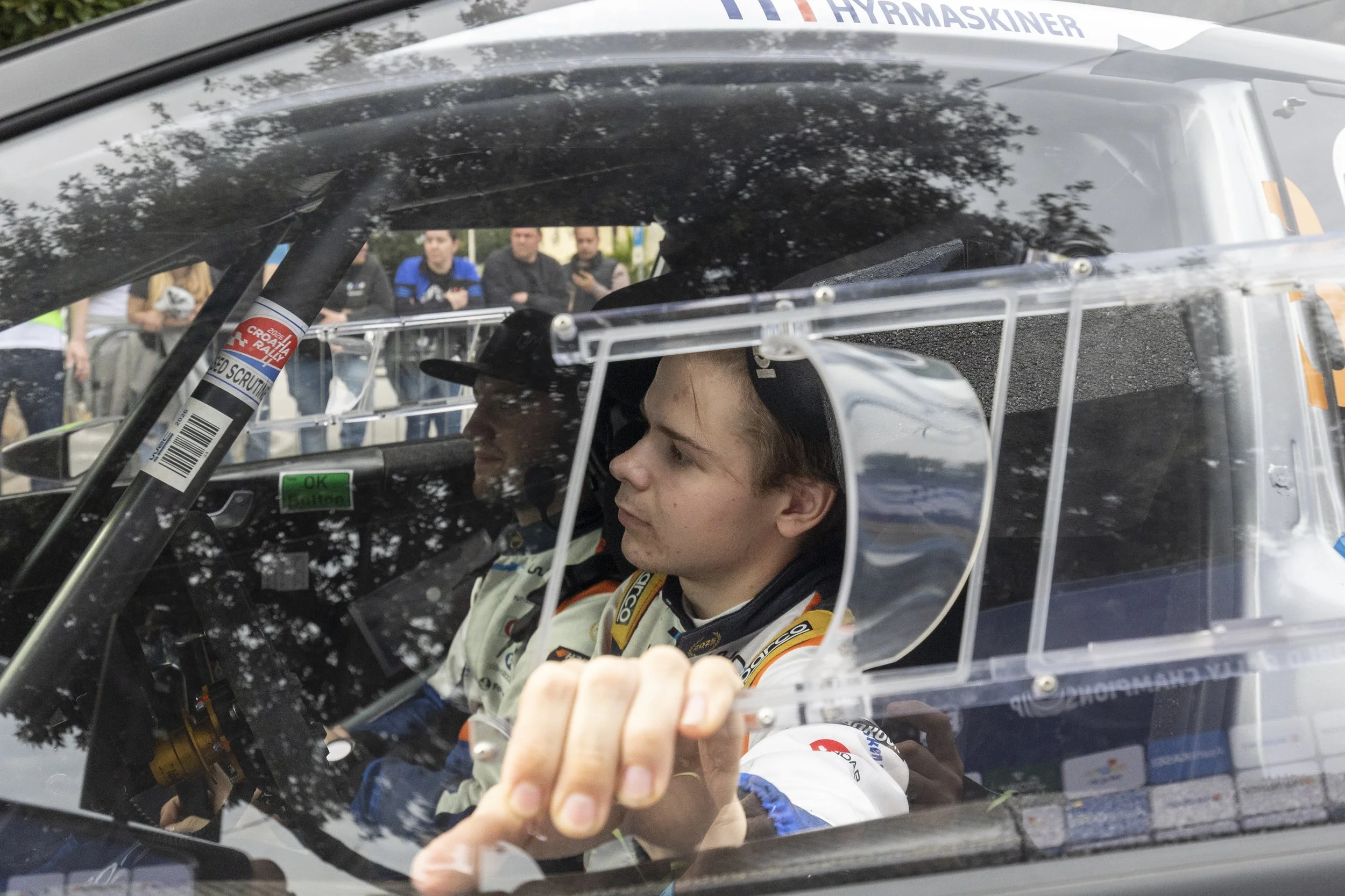 A racing driver and a young girl sitting inside a race car with a cage, with onlookers in the background.