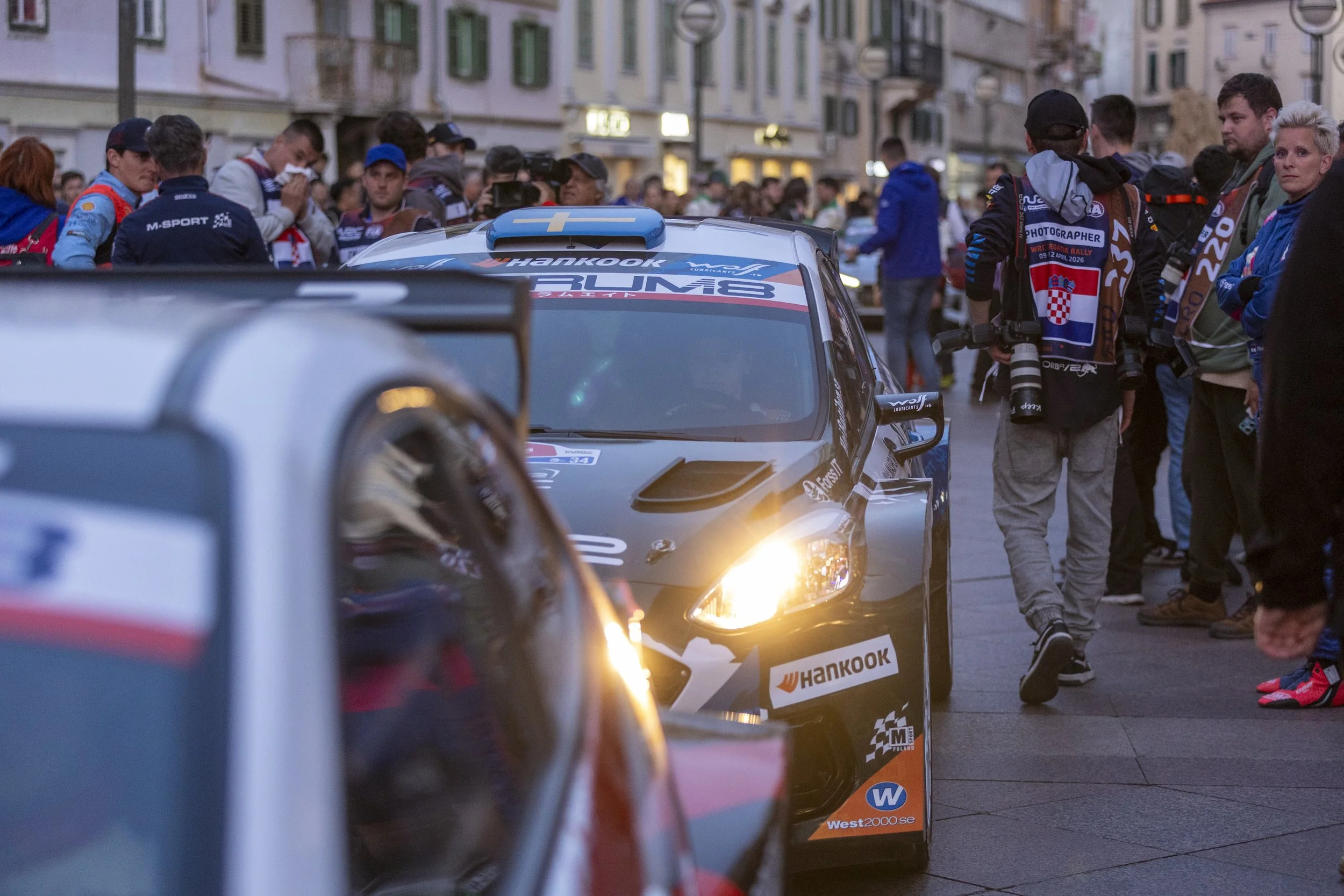A crowded street scene with race cars and people, including photographers and spectators, during a motorsport event. One racing car with sponsorship logos and a blue fin on top is visible, with a crowd of people around it.
