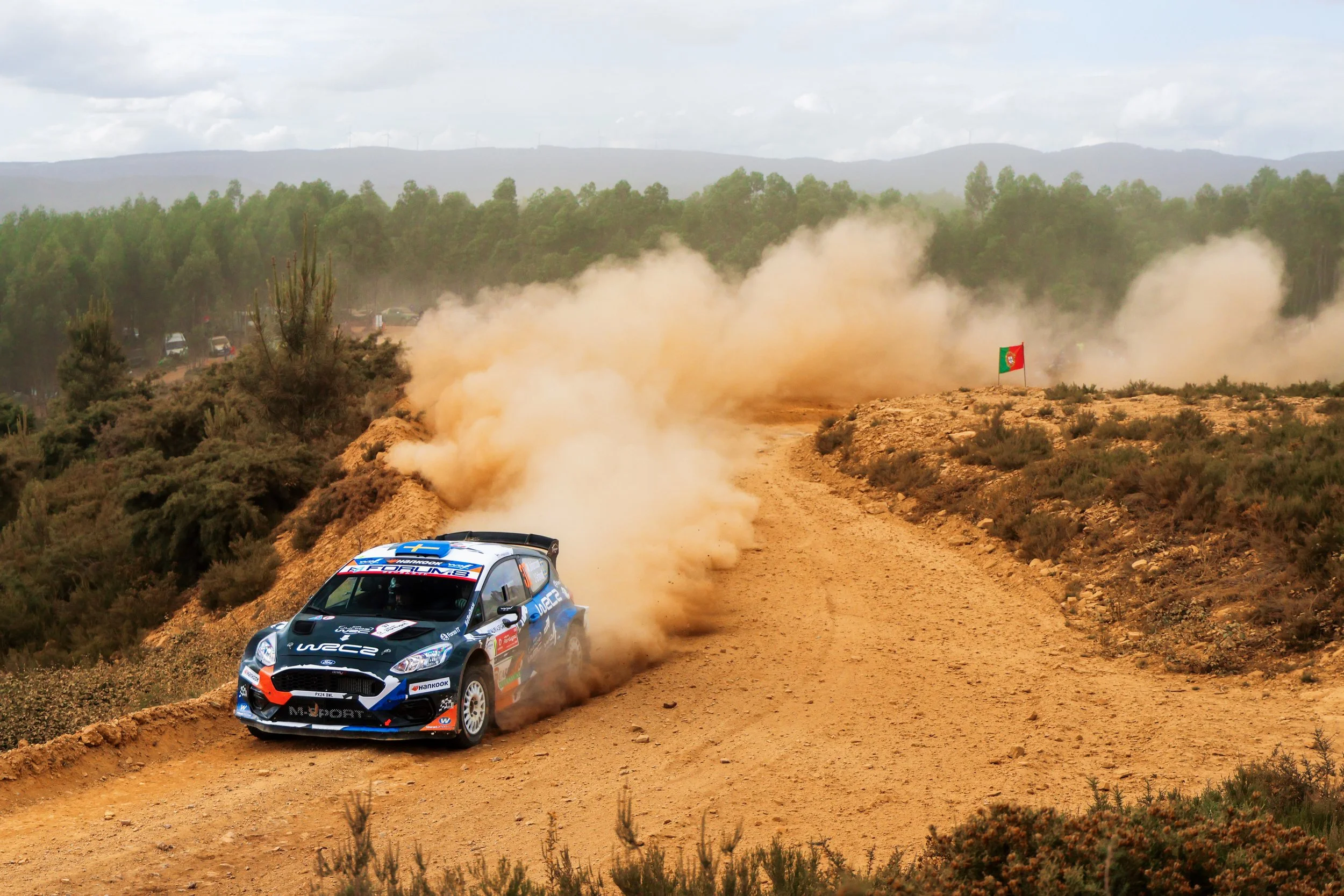 A rally car racing on a dirt road, kicking up dust, with a Portuguese flag in the background and a forested landscape.